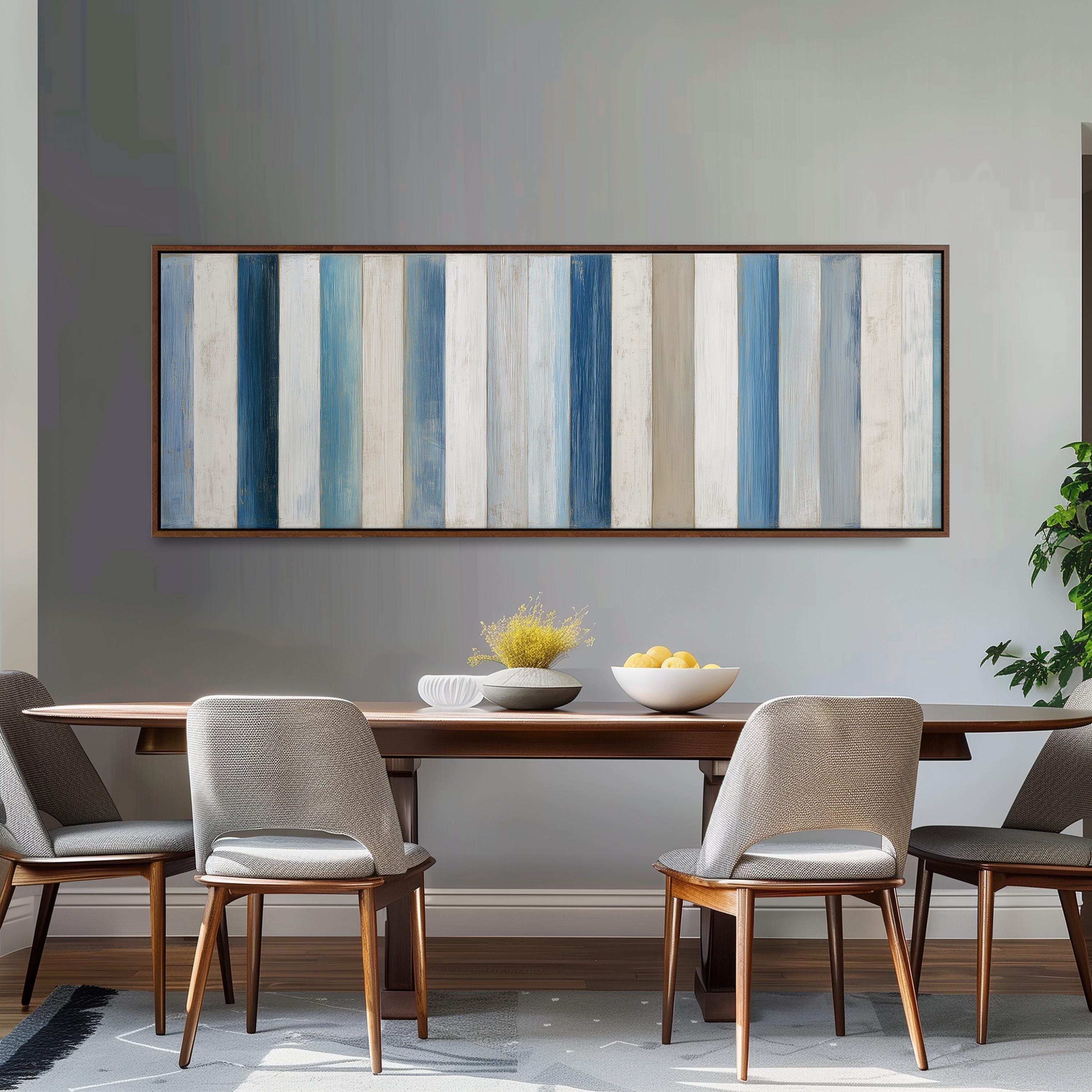 a dining room with a wooden table and four chairs. Above the table, there is a large painting with vertical stripes in various shades of blue and white. The painting is hung on a gray wall. On the table, there is a bowl of fruit and a vase of flowers.