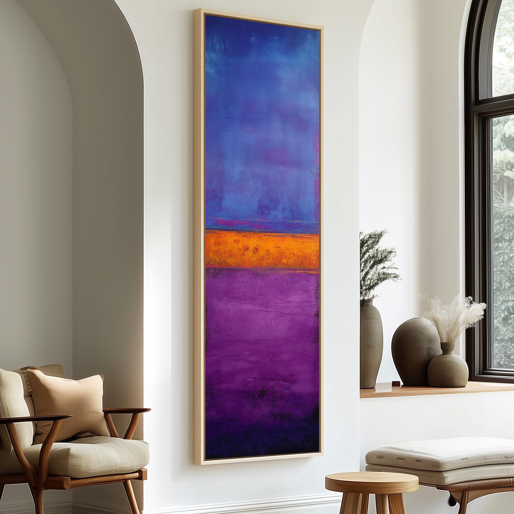 a large abstract painting with a vibrant mix of blue, purple, and orange colors, hanging on a white wall. The painting is framed and appears to be the focal point of the room. In the background, there is a window with a view of trees, and a wooden chair is placed near the painting.