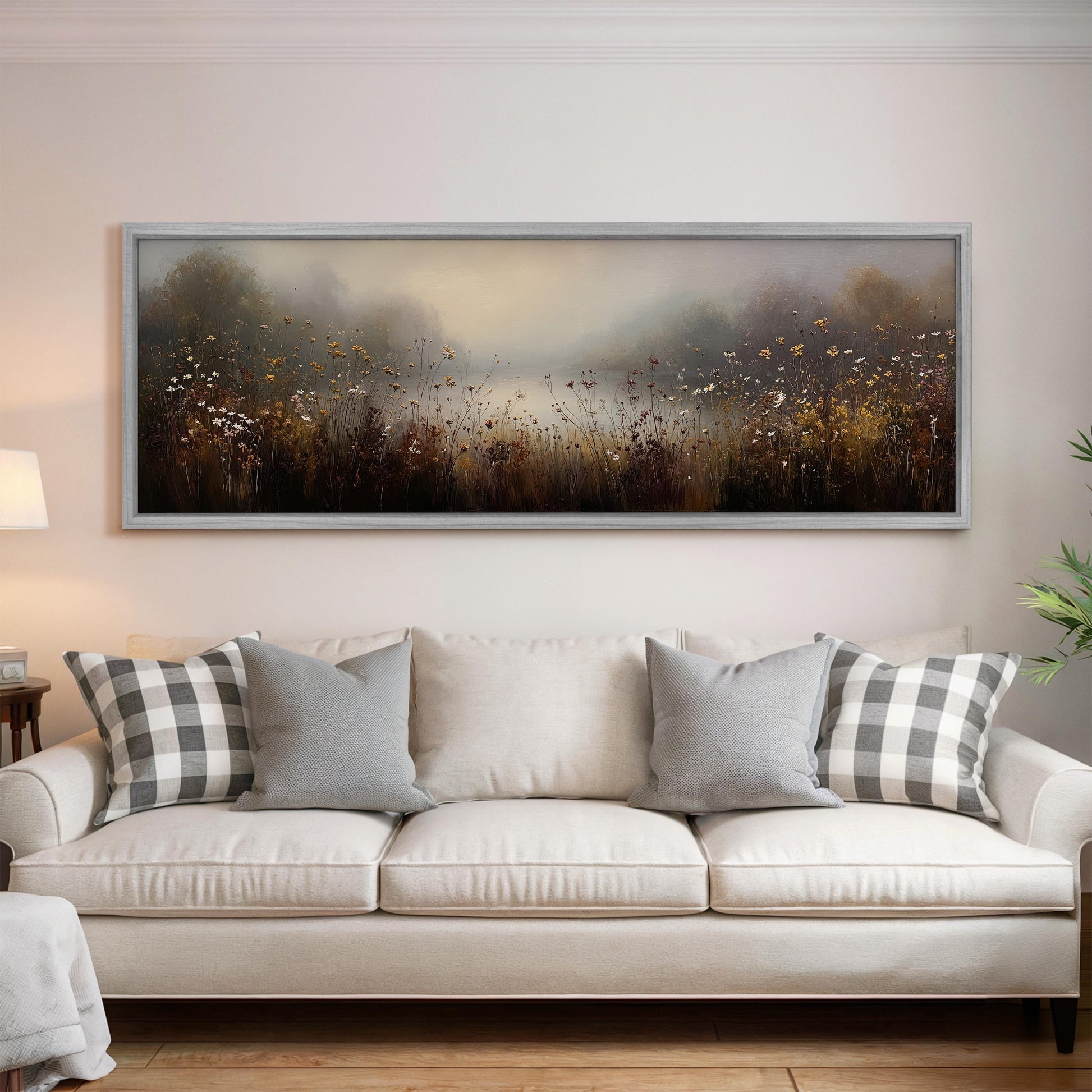 a large, framed painting of a field with flowers hanging on a wall above a white couch with gray and white plaid throw pillows.