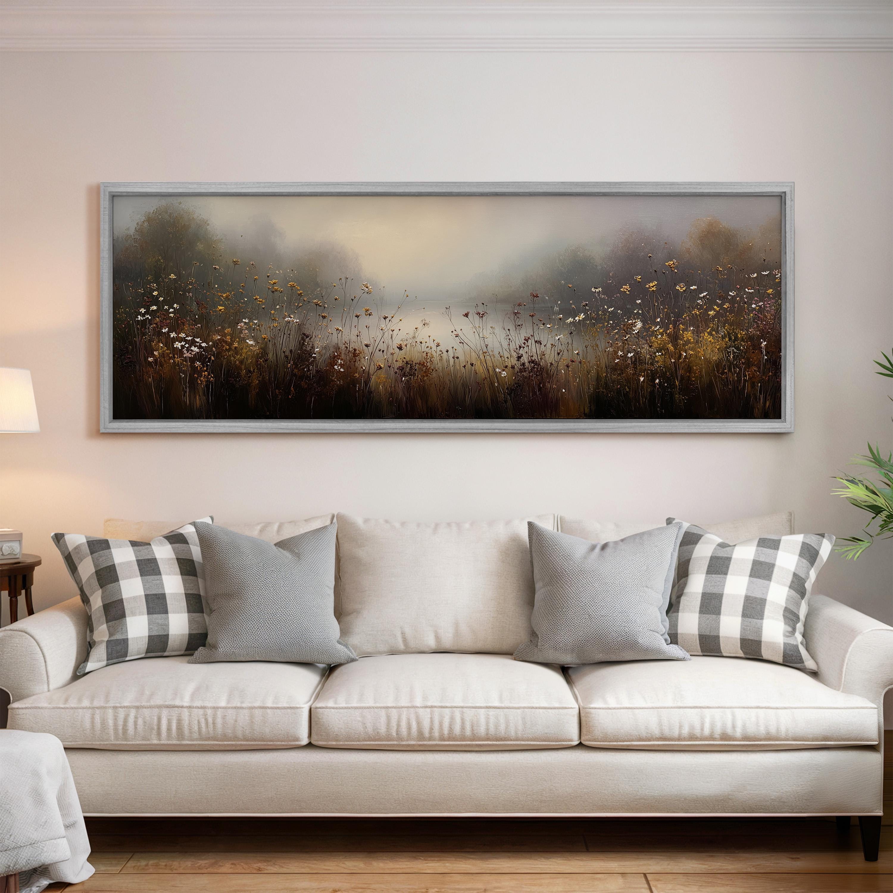 a large, framed painting of a field with flowers hanging on a wall above a white couch with gray and white plaid throw pillows.