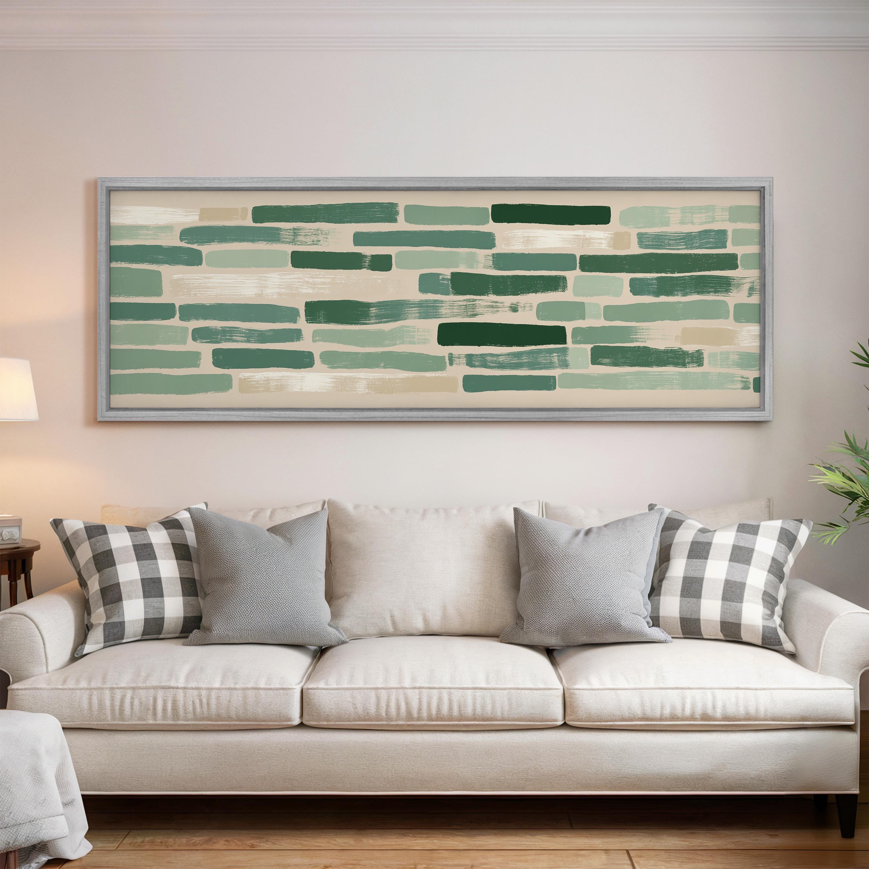 a living room with a white sofa adorned with gray and white patterned pillows. Above the sofa, there is a large framed artwork depicting a pattern of green and white horizontal lines.