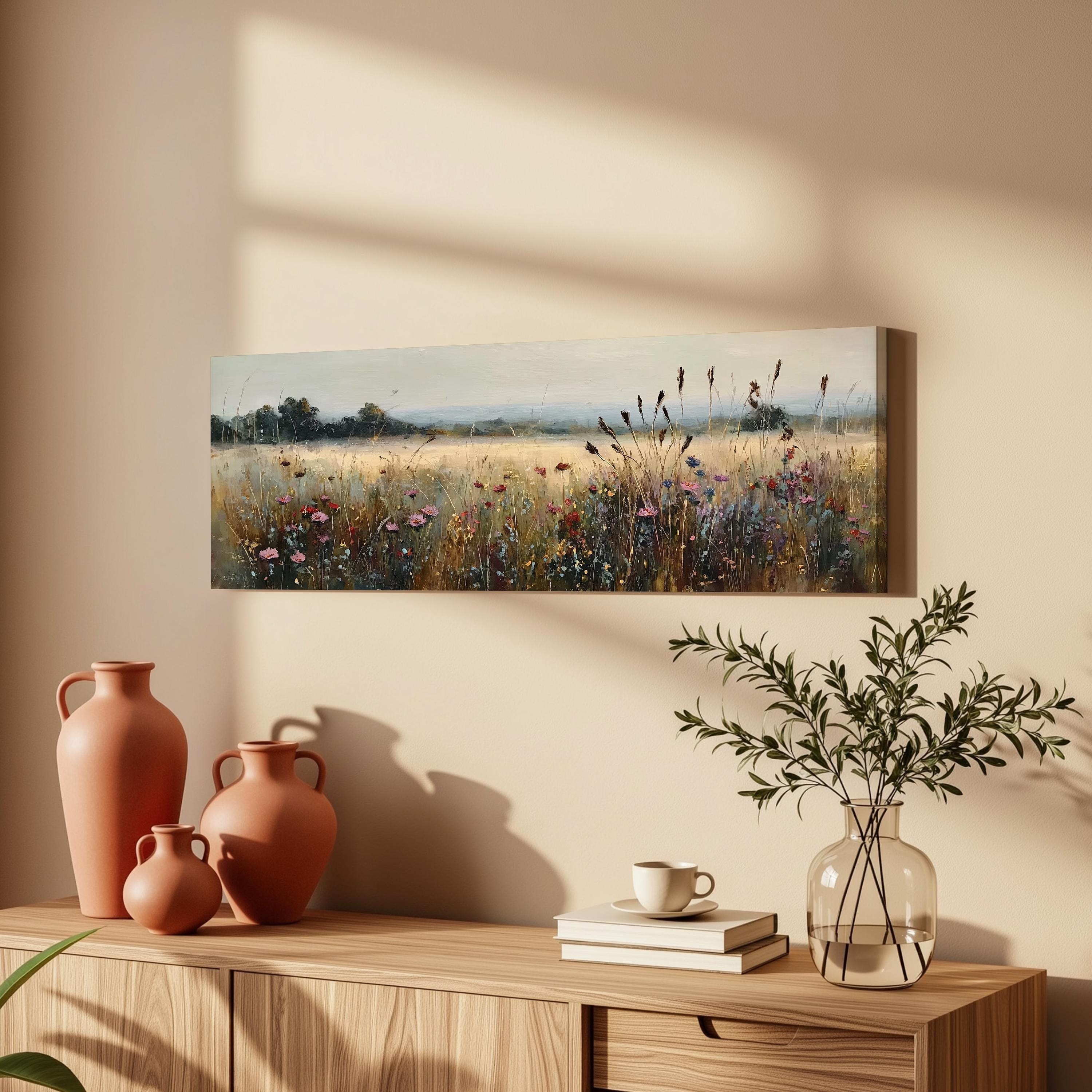a wooden cabinet with a painting of a field of flowers hanging on the wall above it. The cabinet also has three orange vases and a glass vase with greenery on top of it.