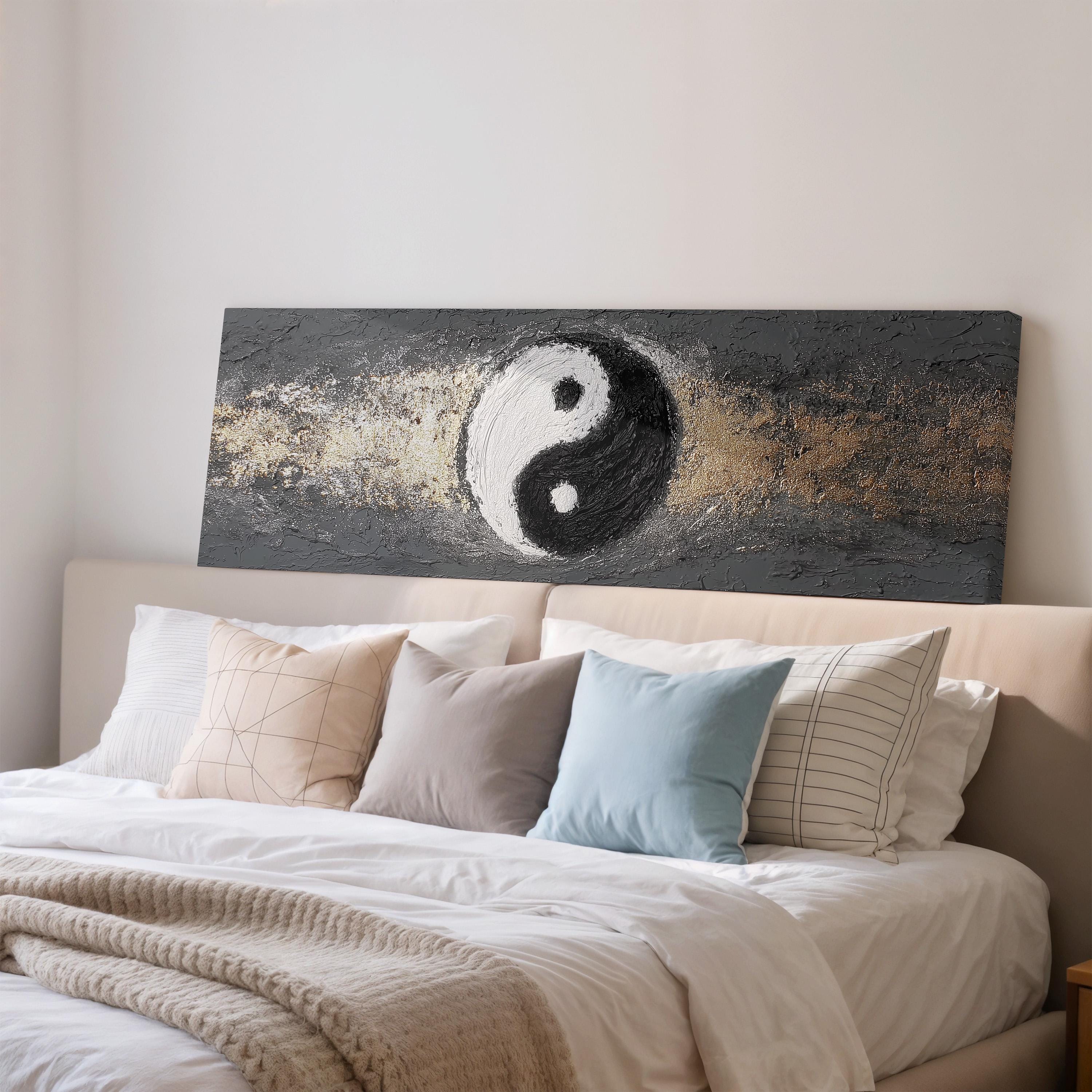 a large bed with a white comforter and various pillows, including a blue pillow and a striped pillow. Above the bed, there is a large canvas print depicting a yin and yang symbol, which is a common symbol in Eastern spiritual and philosophical traditions.
