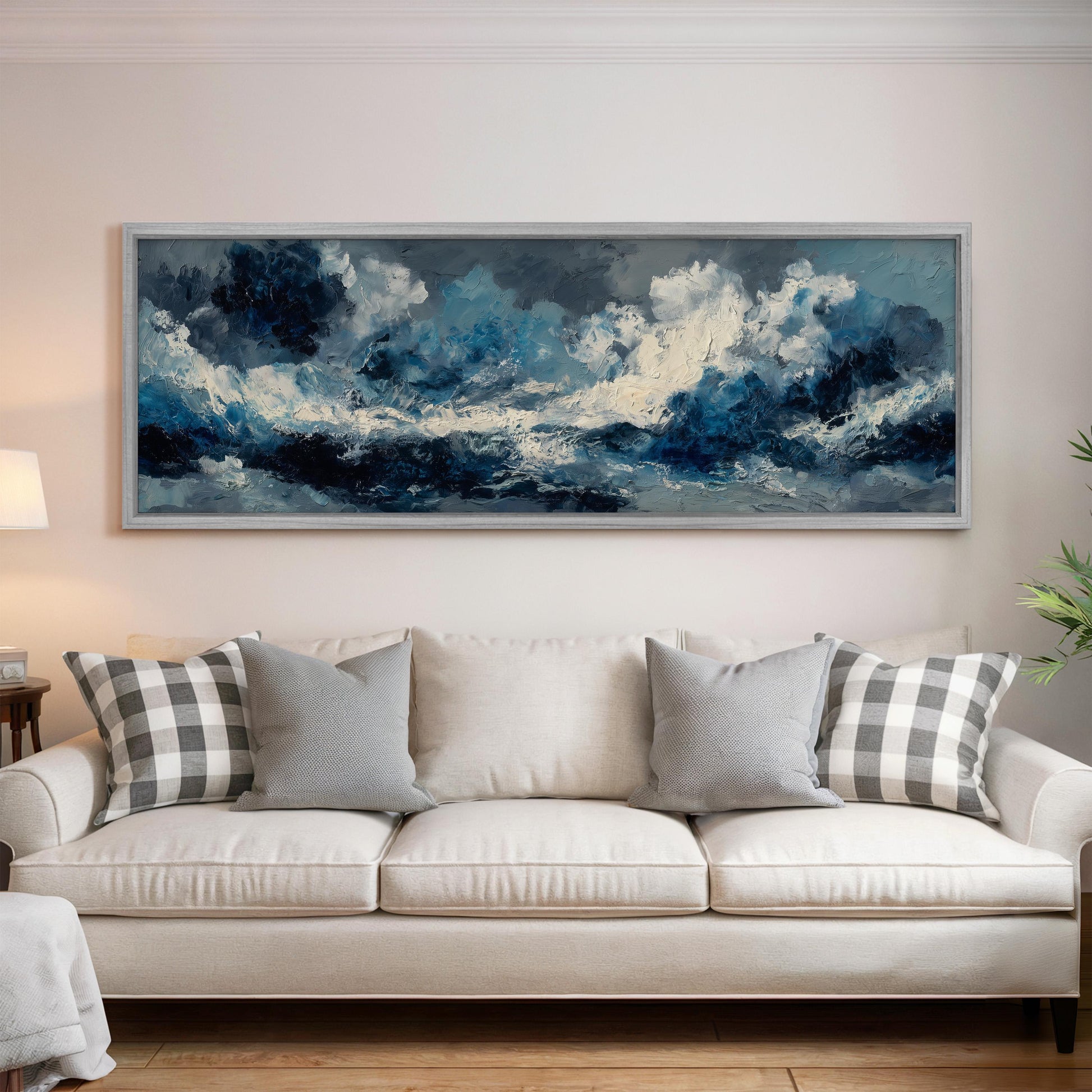 a large, framed painting of a stormy ocean scene hanging on a wall above a white sofa with gray and white patterned throw pillows.