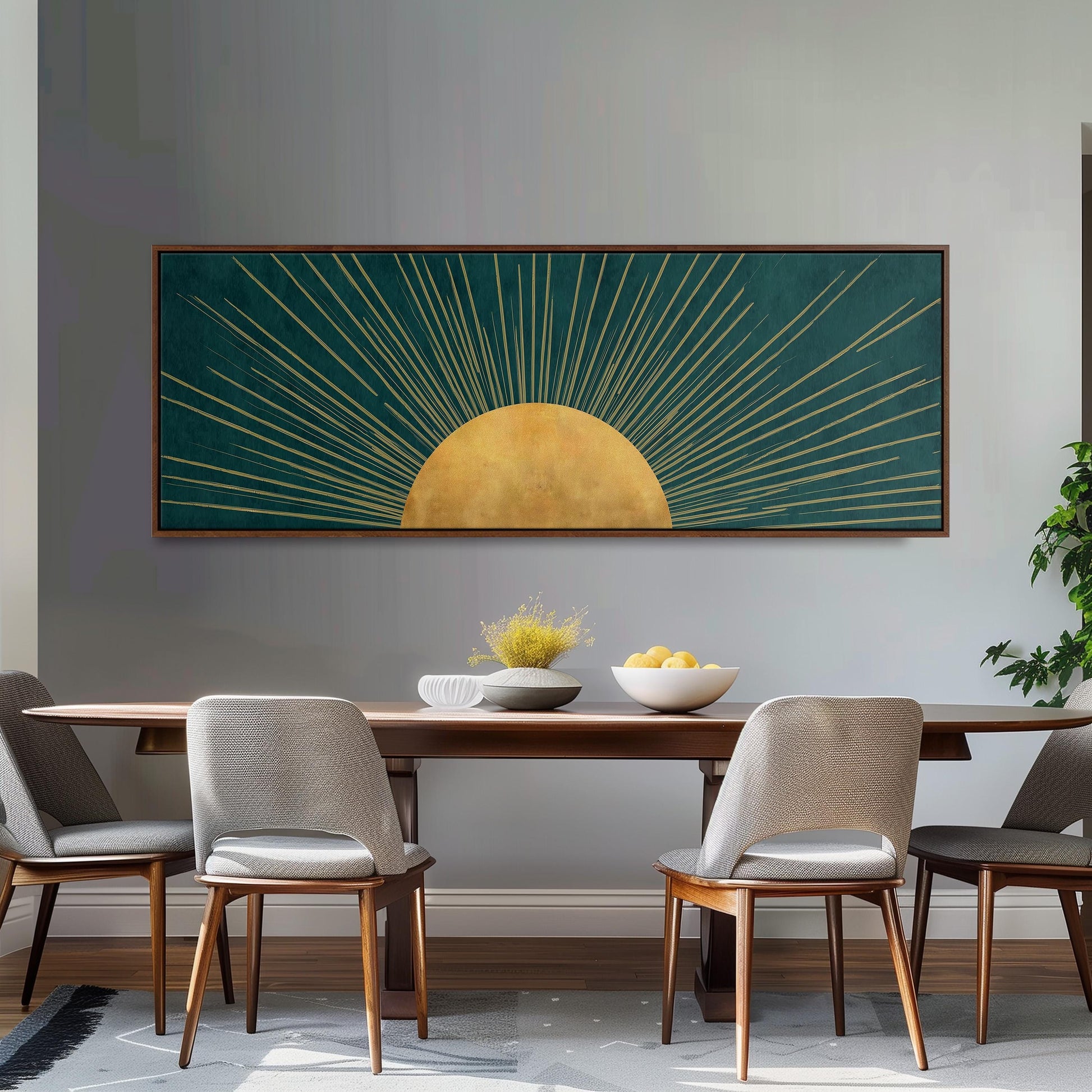 a dining room with a wooden table and four chairs. Above the table, there is a large framed artwork depicting a sun with rays radiating from it. The artwork is set against a gray wall, and the table is adorned with a bowl of fruit and a vase of flowers.