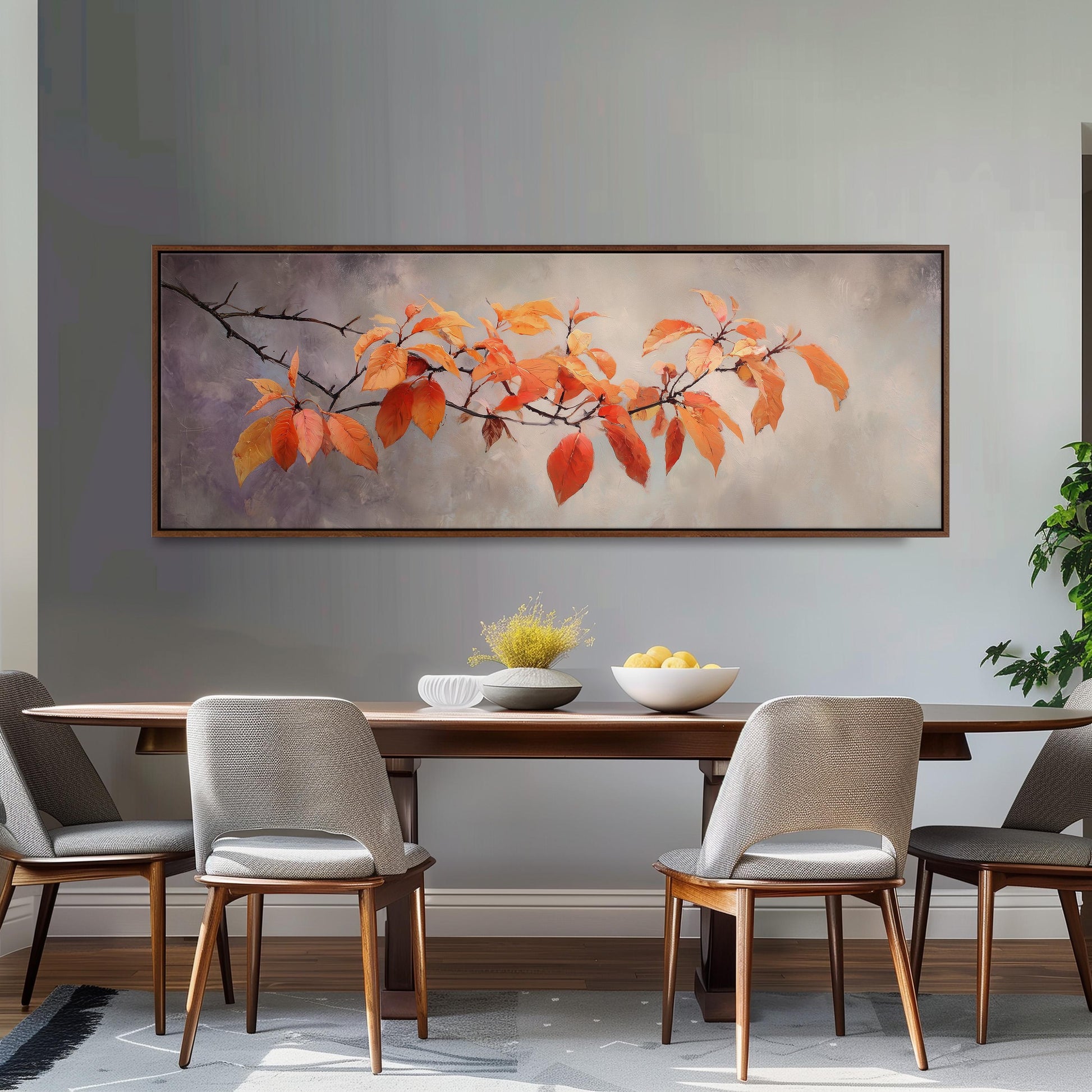 a dining room with a wooden table and four chairs. Above the table, there is a large painting of orange leaves on a branch. The painting is framed and hangs on the wall. On the table, there is a bowl of fruit and a vase of flowers.