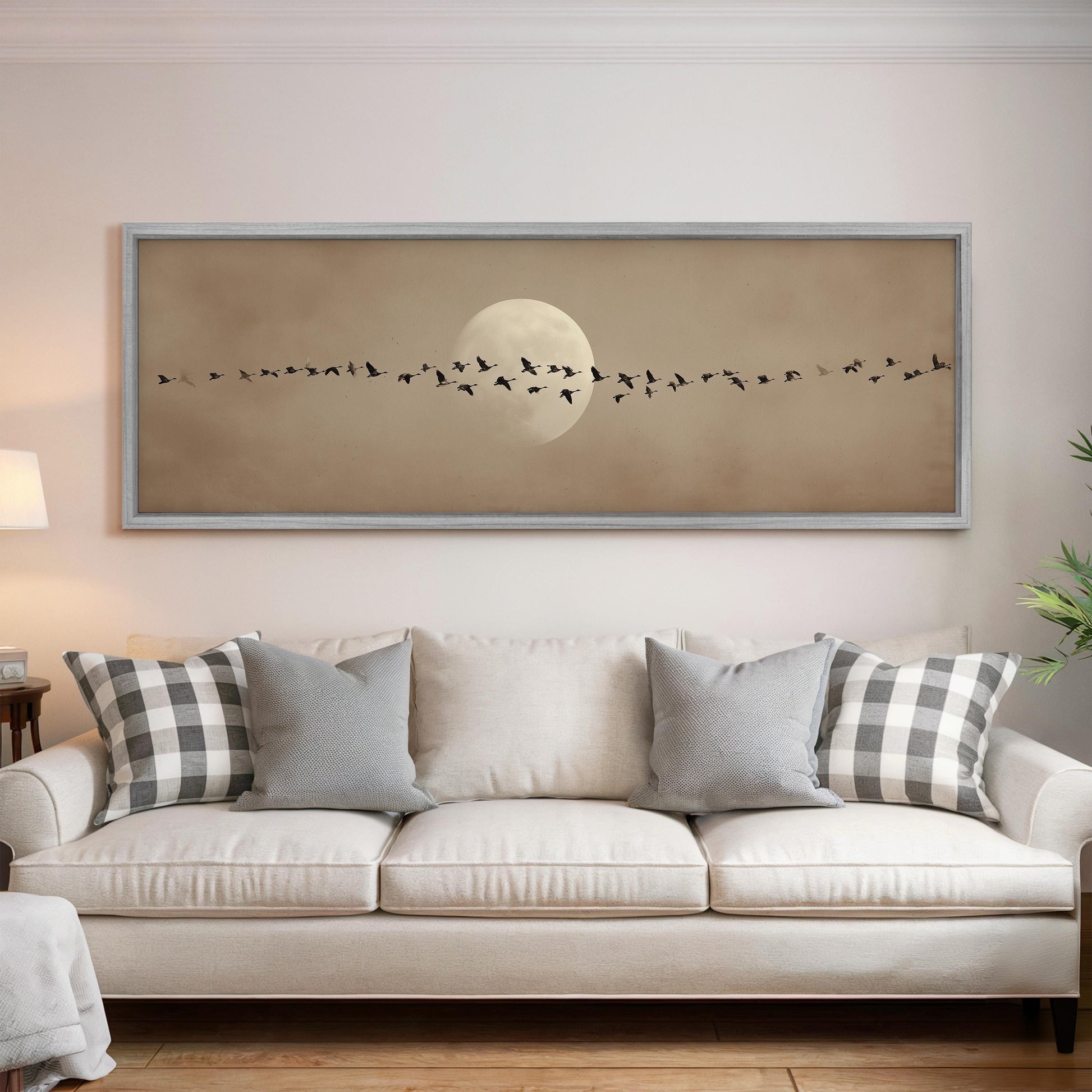 a large framed picture of a flock of birds flying across a moonlit sky, hanging on a wall above a white couch with patterned throw pillows.