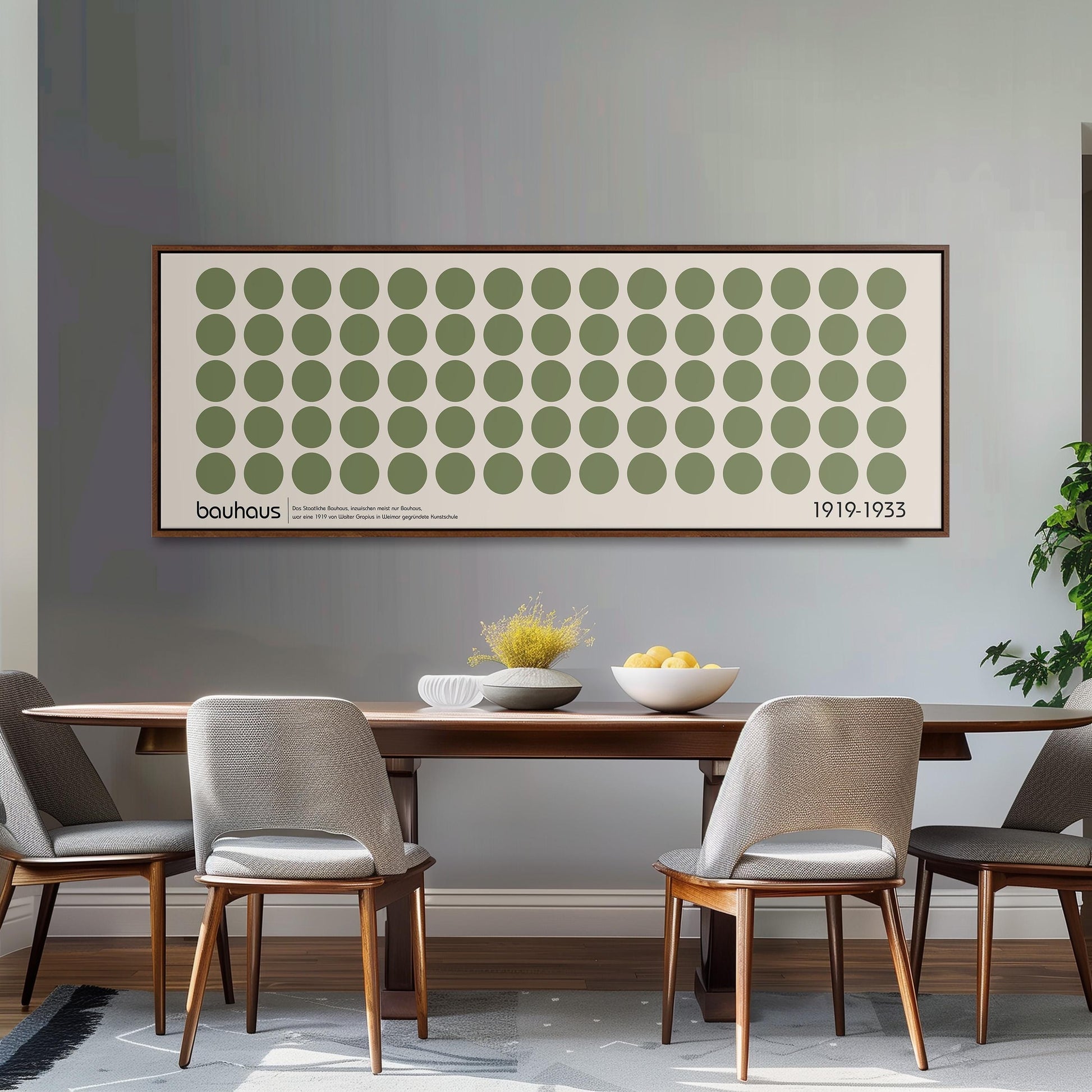 a dining room with a wooden table and four chairs. Above the table, there is a large framed poster with a repeating pattern of green circles and white dots. The poster is hanging on a gray wall. On the table, there is a bowl of fruit and a vase of flowers.