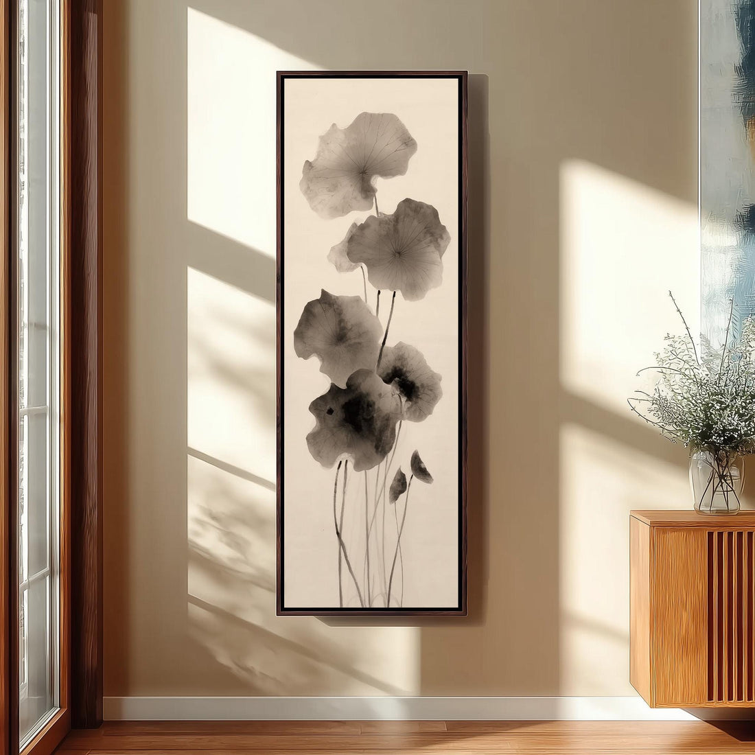 a minimalist living room with a large framed artwork of black and white flowers hanging on the wall. The room is furnished with a chair, a table, and a window with a plant on the sill.