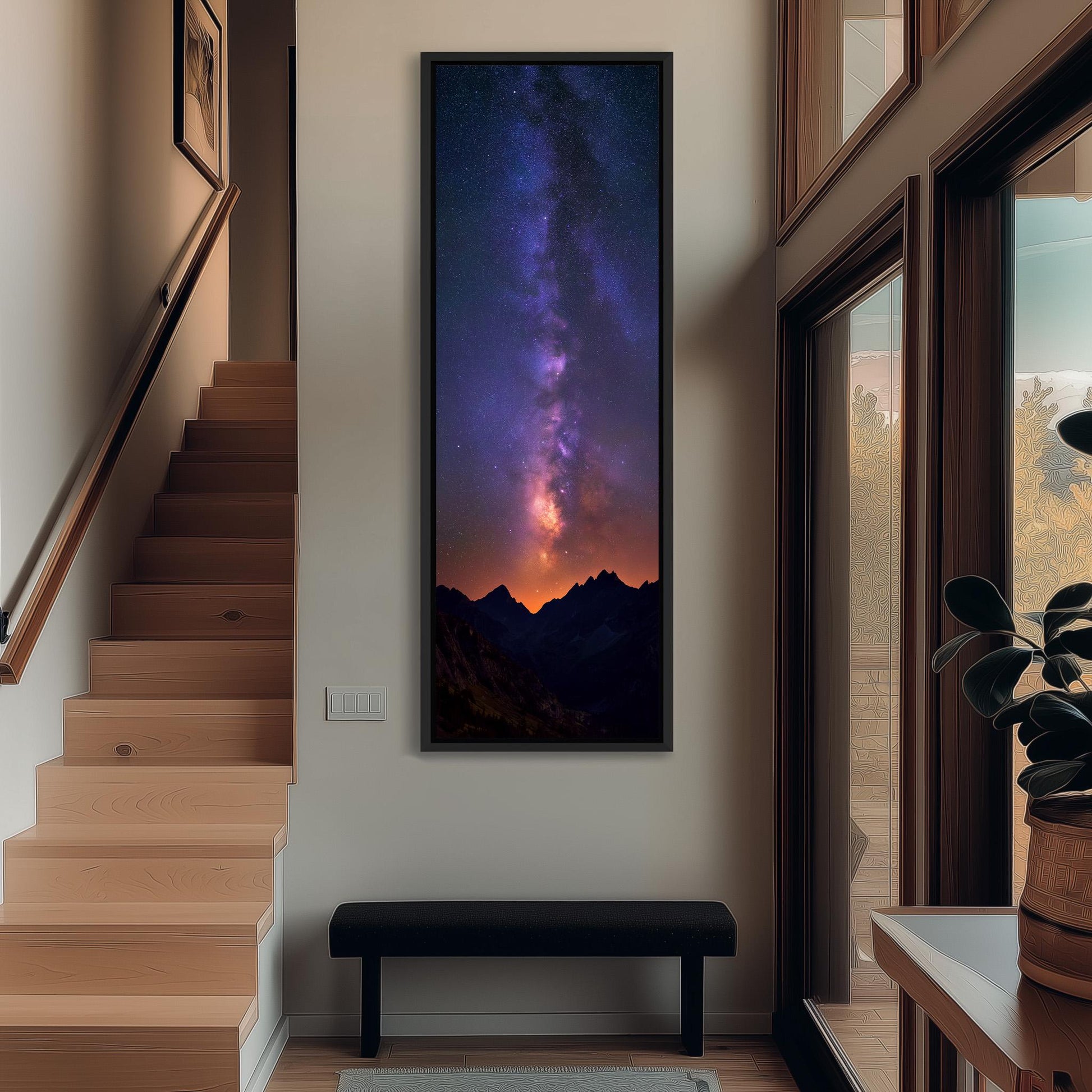 a large, framed picture of a night sky with a galaxy, hanging on a wall in a hallway. The hallway has a wooden staircase and a bench, and there is a plant on the right side of the image.