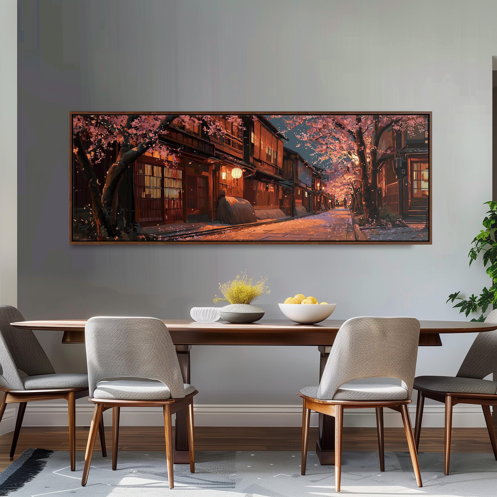 a large, framed painting of a serene street scene with cherry blossoms and a warm, glowing building at the end of the street. The painting is hung on a wall above a wooden dining table with four chairs. The table is set with a bowl of fruit and a vase of flowers, creating a cozy and inviting atmosphere.