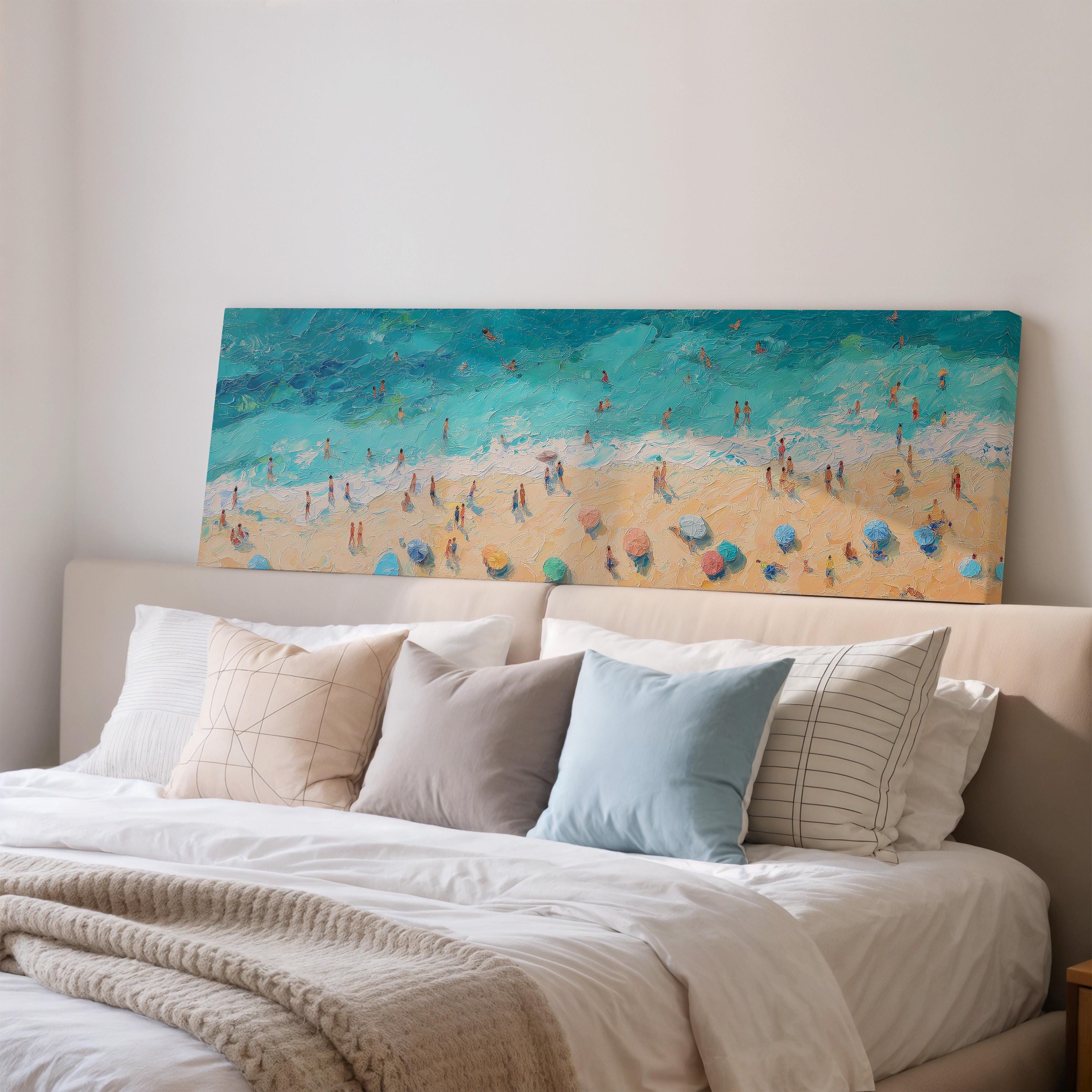 a large painting of a beach scene with people and umbrellas, hanging above a bed with white pillows and a gray blanket.