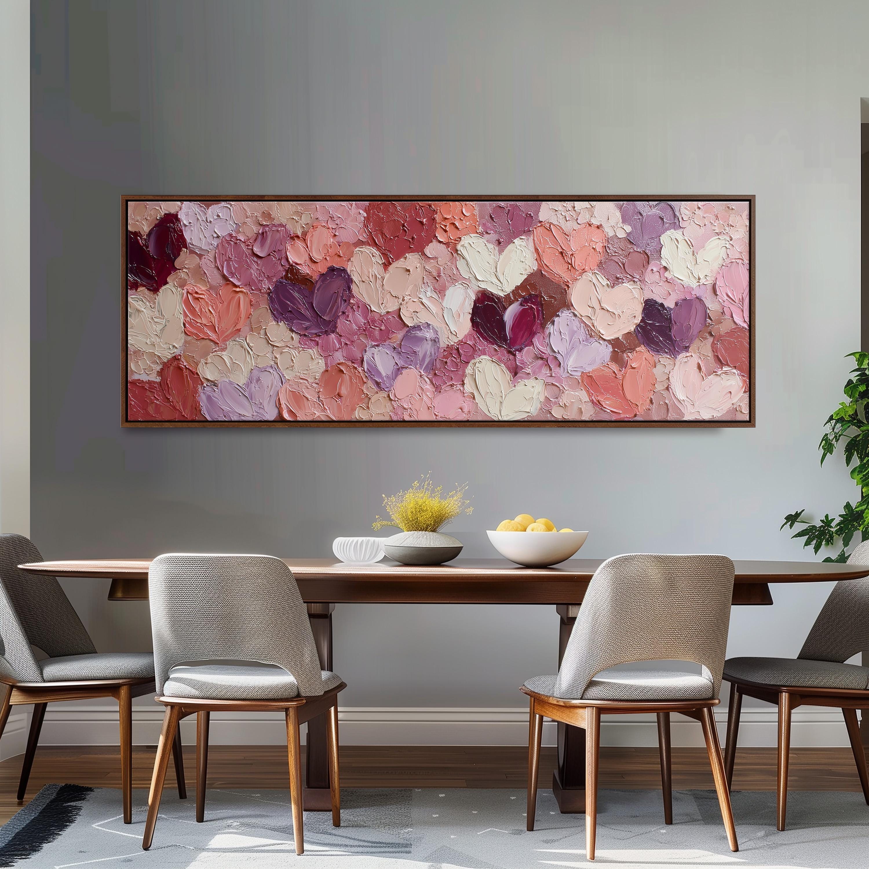 a dining room with a wooden table and four chairs. Above the table, there is a large painting of hearts in various shades of pink and purple. The painting is framed and hangs on the wall above the table.