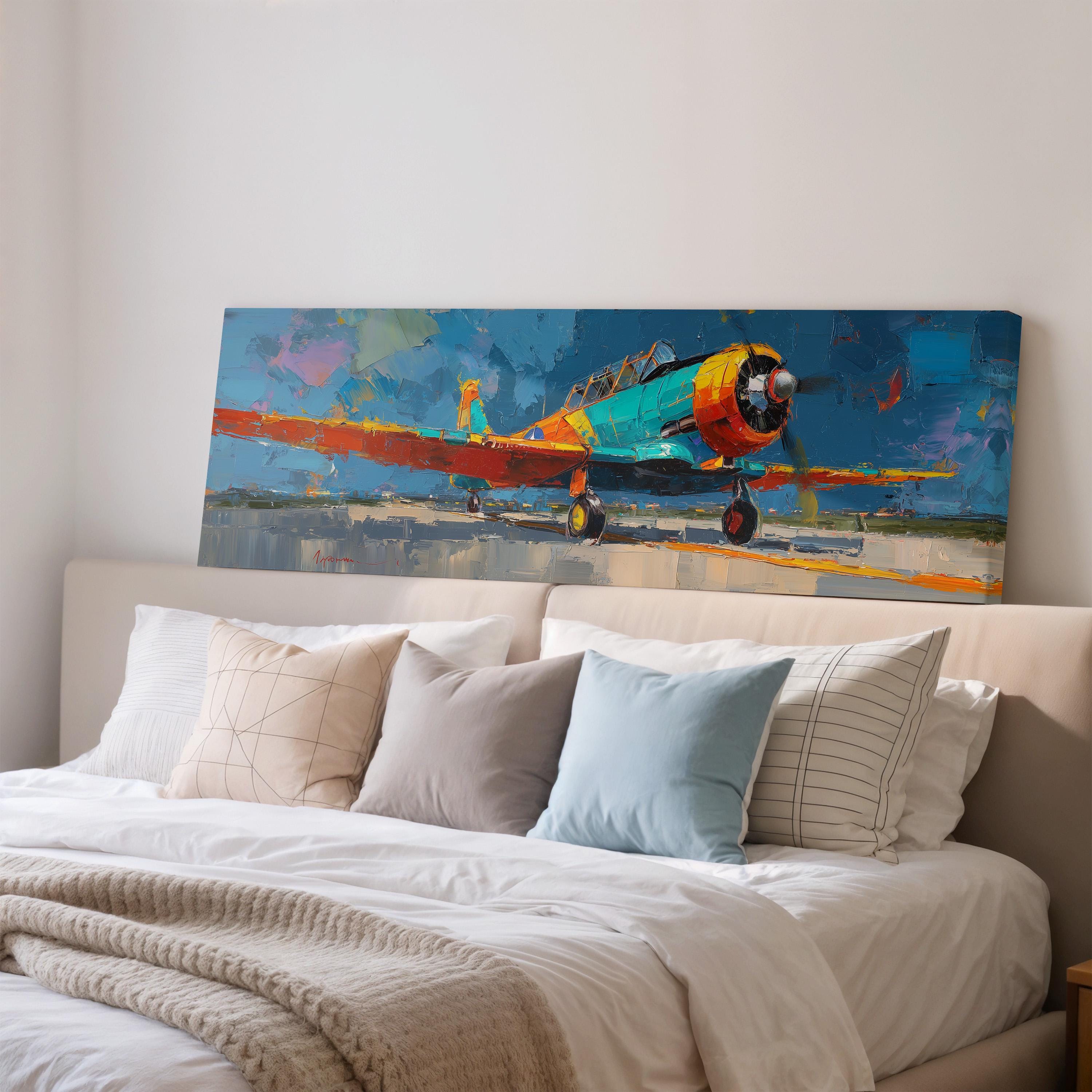 a large painting of a colorful propeller plane hanging on a wall above a bed with white pillows and a gray blanket.
