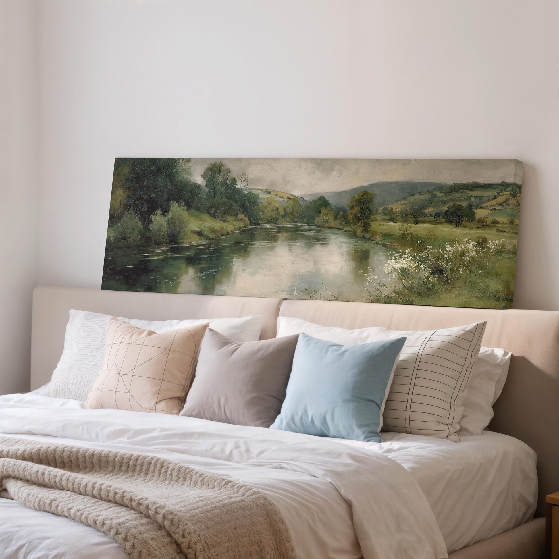 a large painting of a serene landscape with a river and trees, hanging above a bed with white bedding and various pillows.