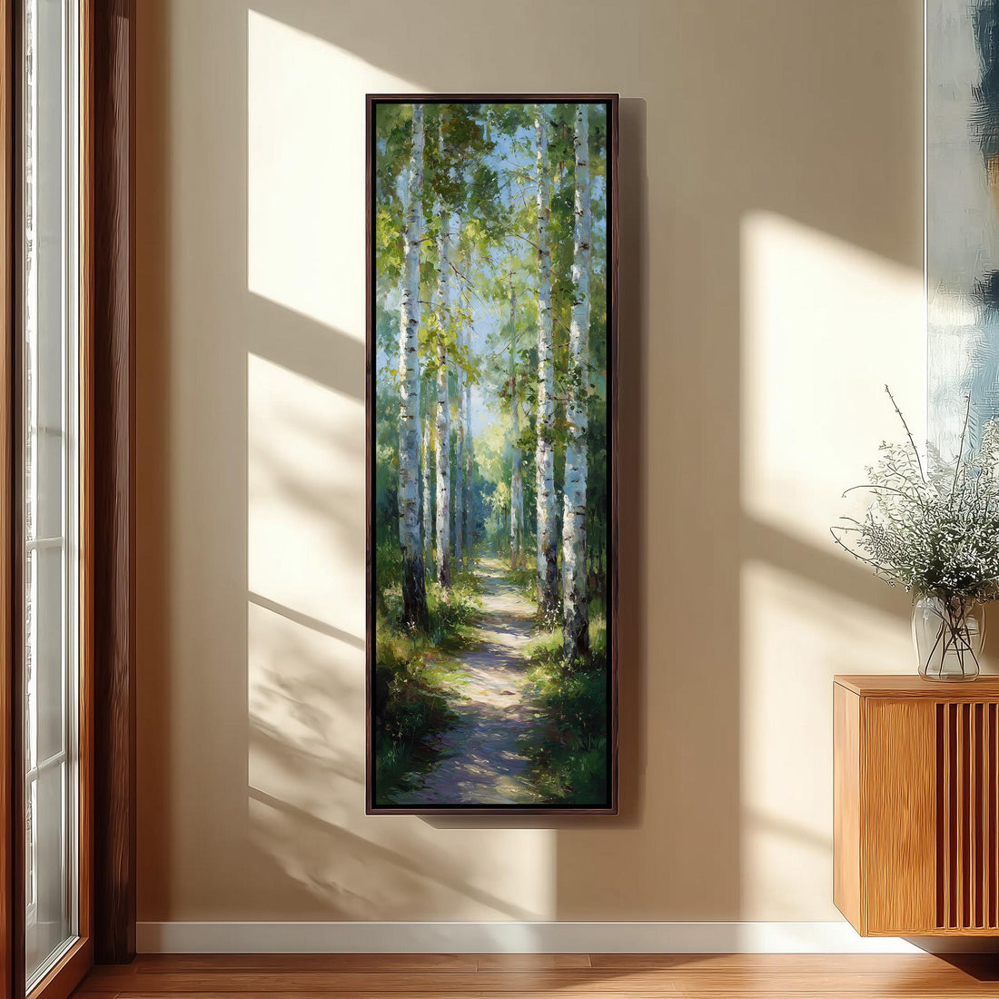 a large painting of a forest path with tall trees and a clear blue sky, hanging on a wall in a room with a wooden floor and a window.