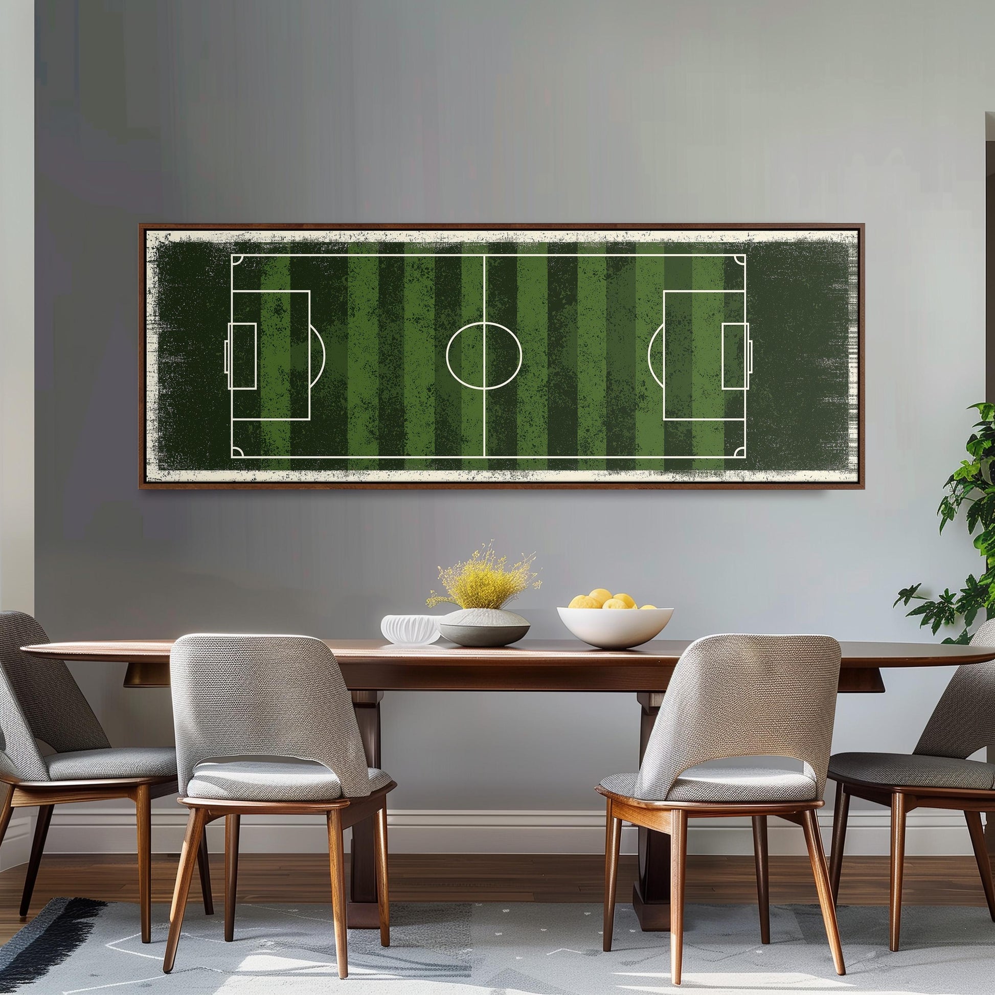 a dining room with a wooden table and four chairs. Above the table, there is a large framed picture of a soccer field with green and white stripes. The table is set with a bowl of fruit and a vase of flowers.