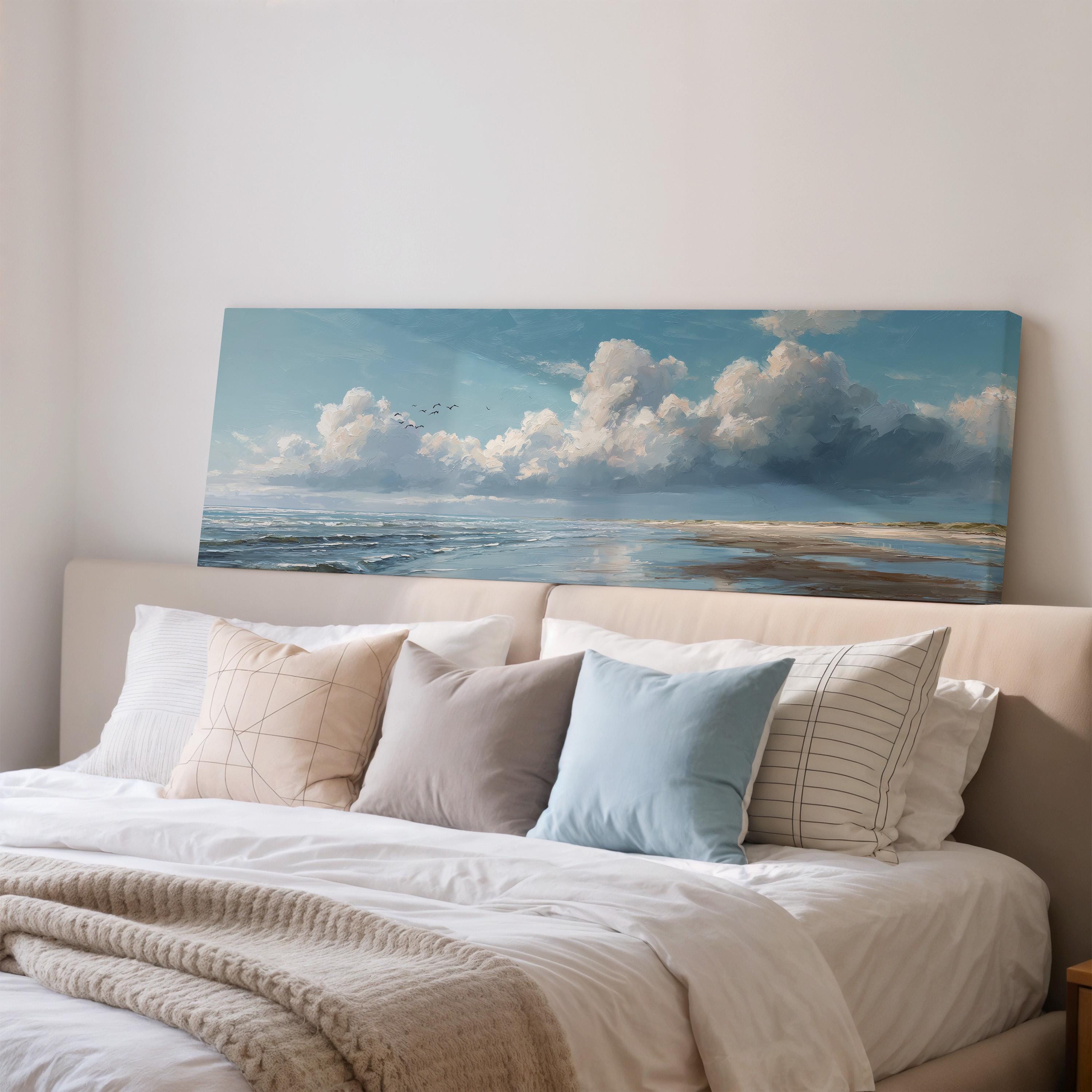 a large painting of a serene ocean landscape with a cloudy sky above, hanging above a bed with white pillows and a gray blanket.
