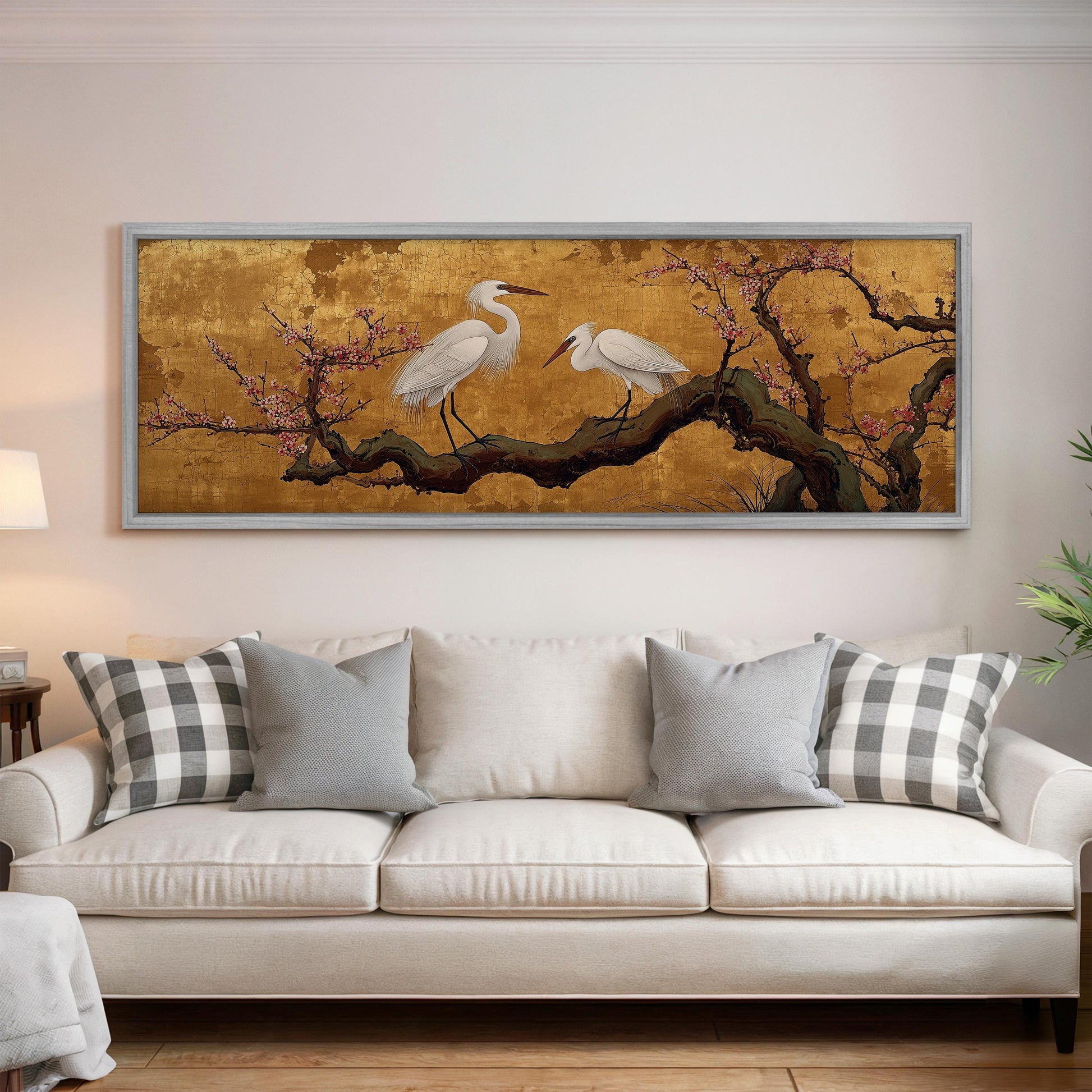 a large, framed painting of two white egrets perched on a branch with pink flowers, hanging on a wall above a white couch with gray and white plaid throw pillows.