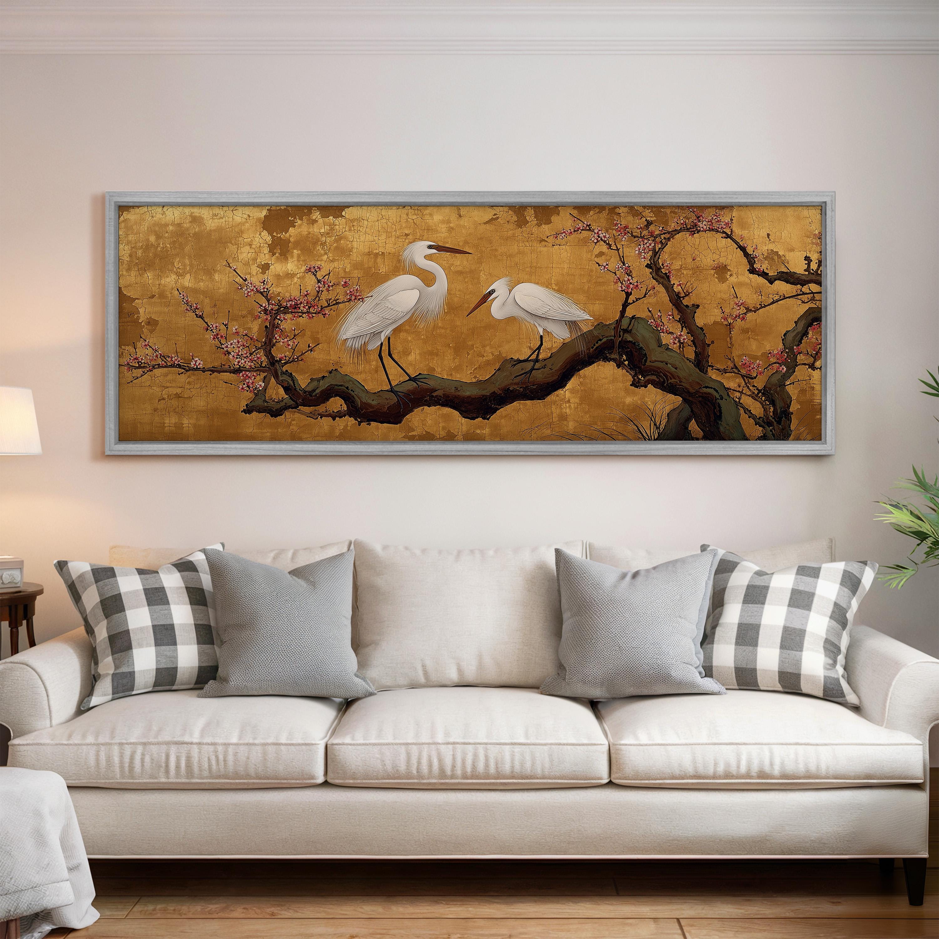 a large, framed painting of two white egrets perched on a branch with pink flowers, hanging on a wall above a white couch with gray and white plaid throw pillows.