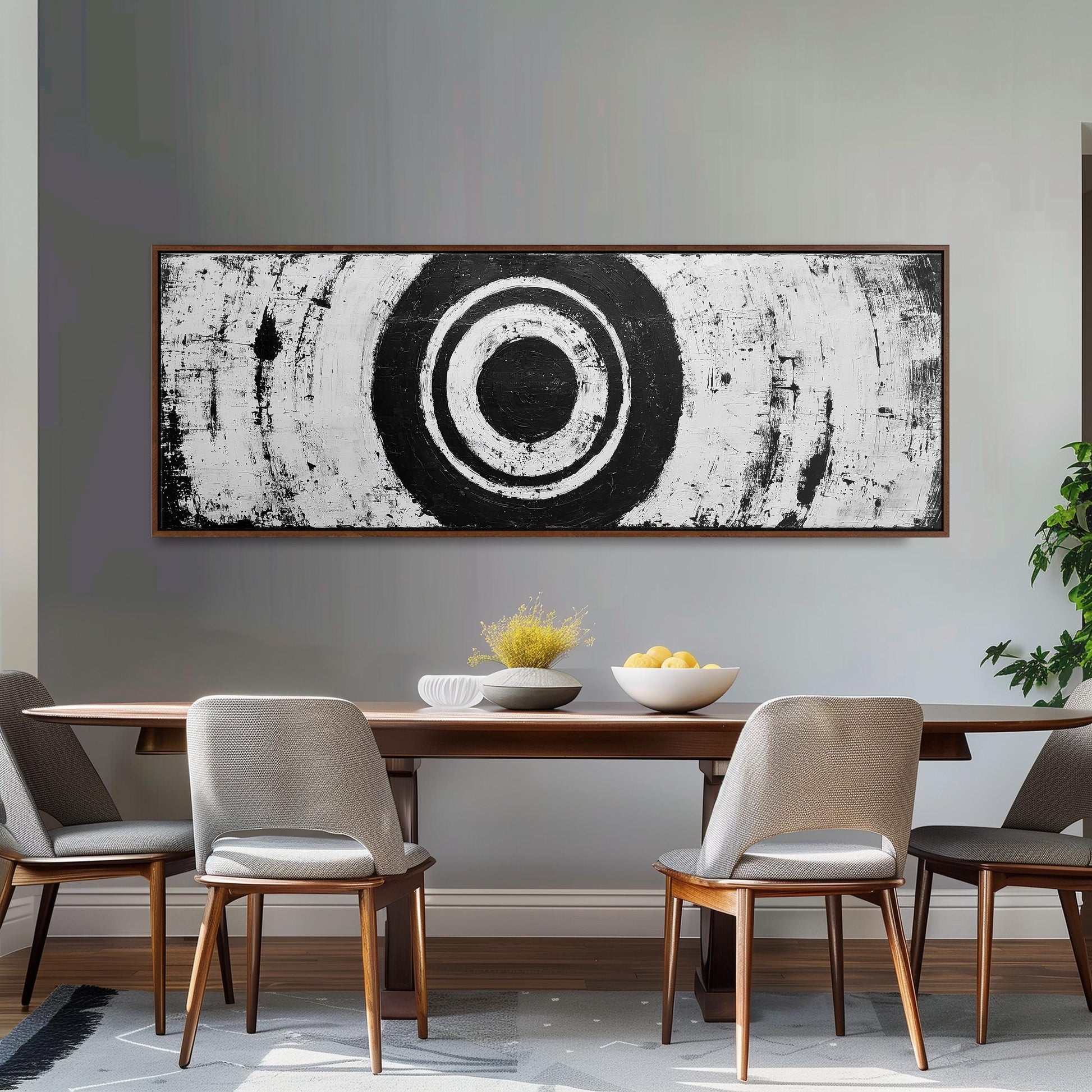 a dining room with a wooden table and four chairs. Above the table, there is a large black and white abstract painting depicting a circle. On the table, there is a bowl of fruit and a vase of flowers.