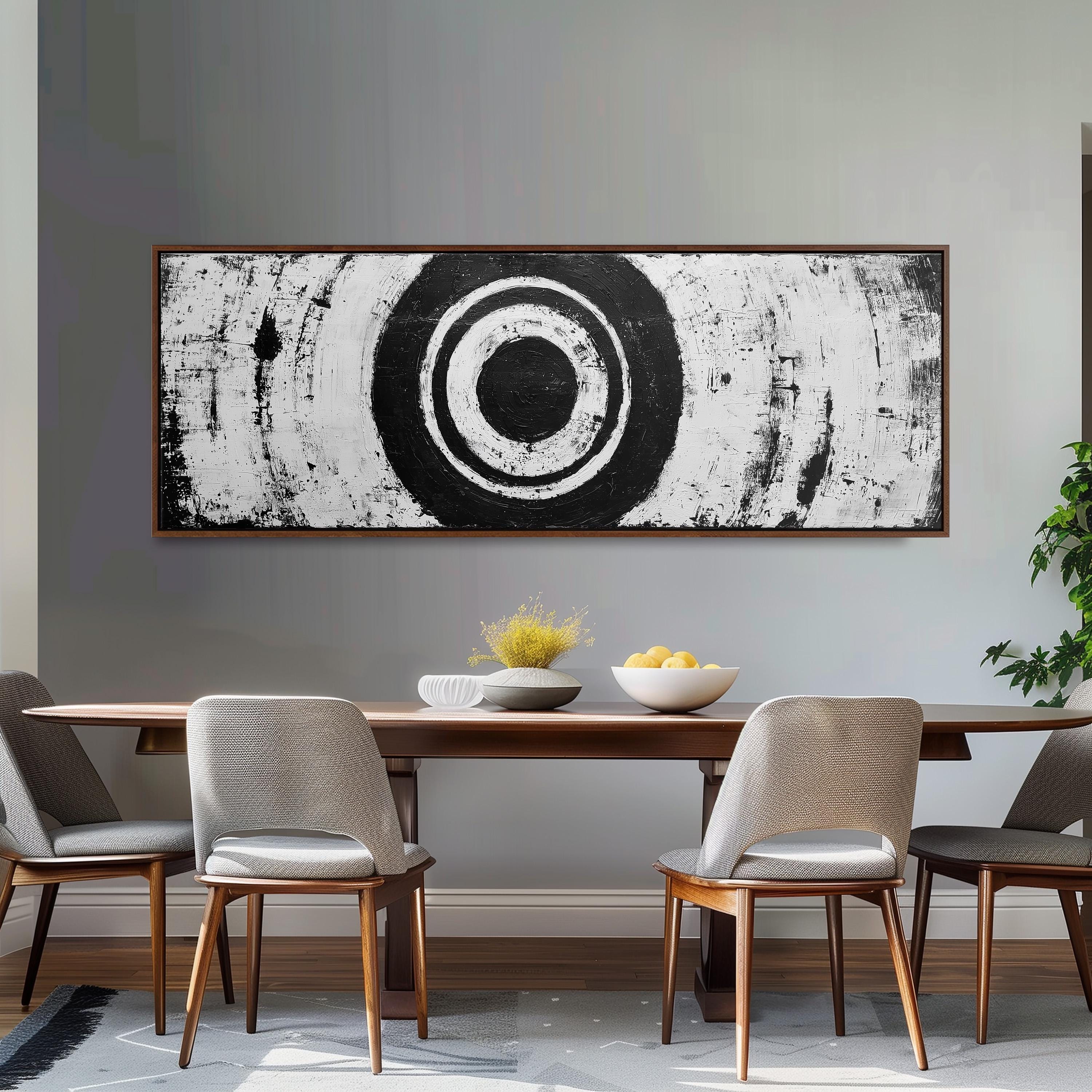 a dining room with a wooden table and four chairs. Above the table, there is a large black and white abstract painting depicting a circle. On the table, there is a bowl of fruit and a vase of flowers.