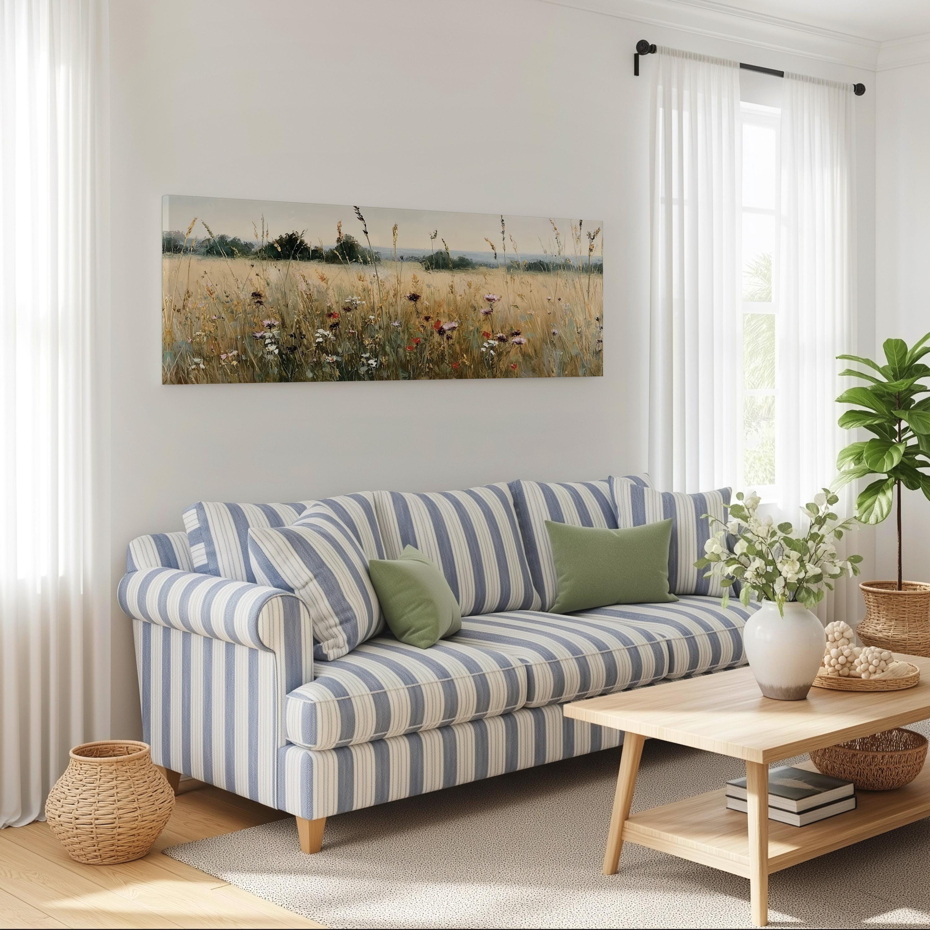 a cozy living room with a striped blue and white sofa, a wooden coffee table, and a large window with white curtains. Above the sofa, there is a painting of a field with tall grass and wildflowers. The room also features a potted plant, a basket, and some books on a shelf.
