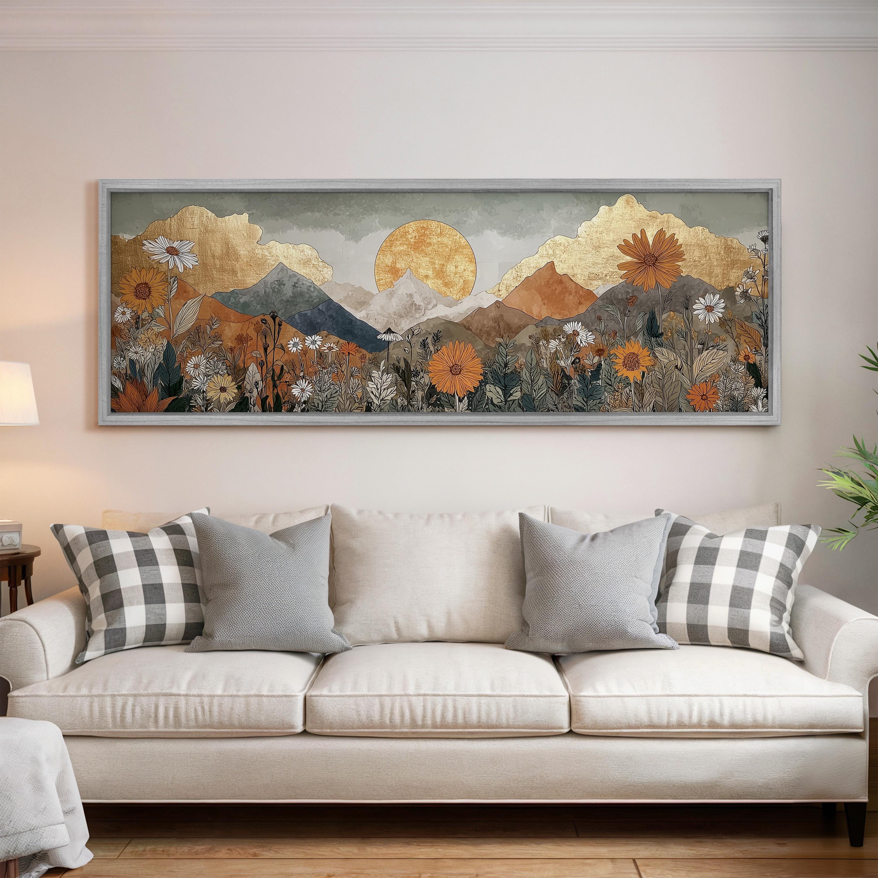 a large, framed painting of a mountainous landscape with a sun in the sky, hanging on a wall above a white couch with decorative pillows.