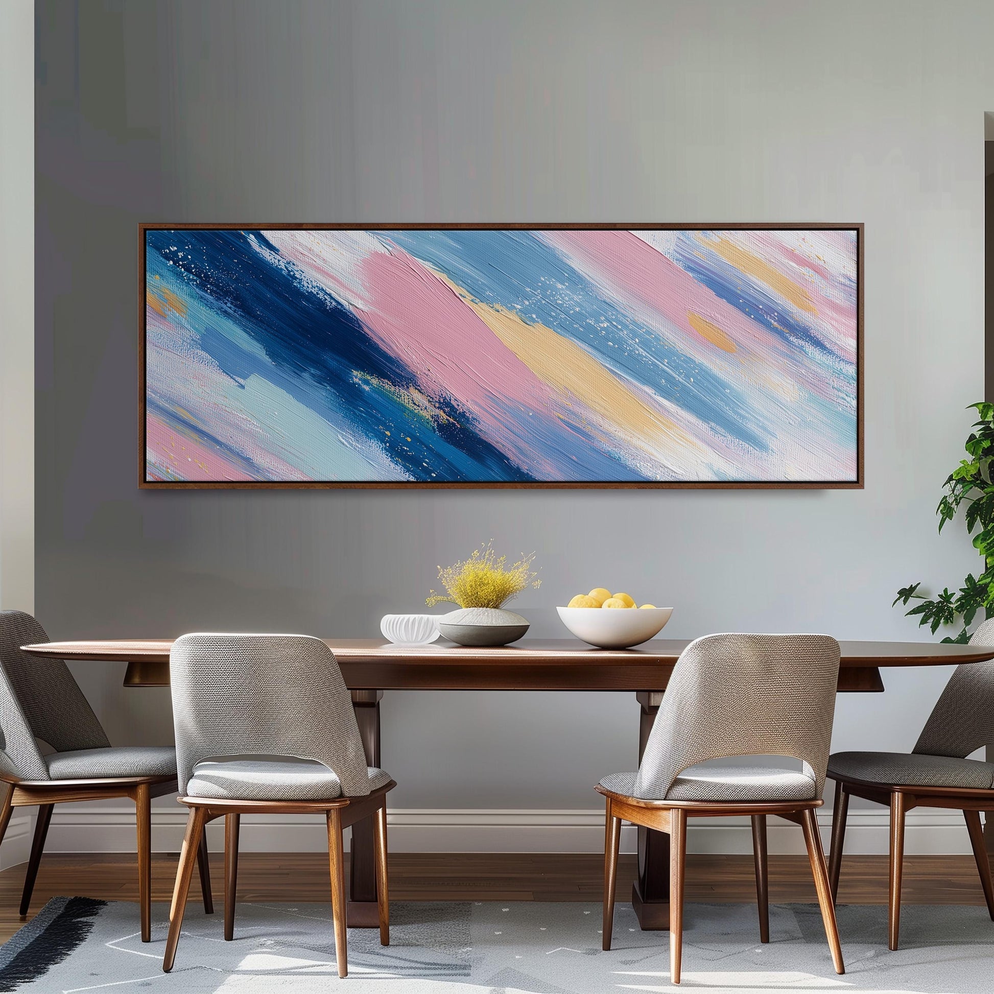 a dining room with a wooden table and four chairs. Above the table, there is a large abstract painting with blue, pink, and yellow brushstrokes. On the table, there is a bowl of fruit and a vase of flowers.