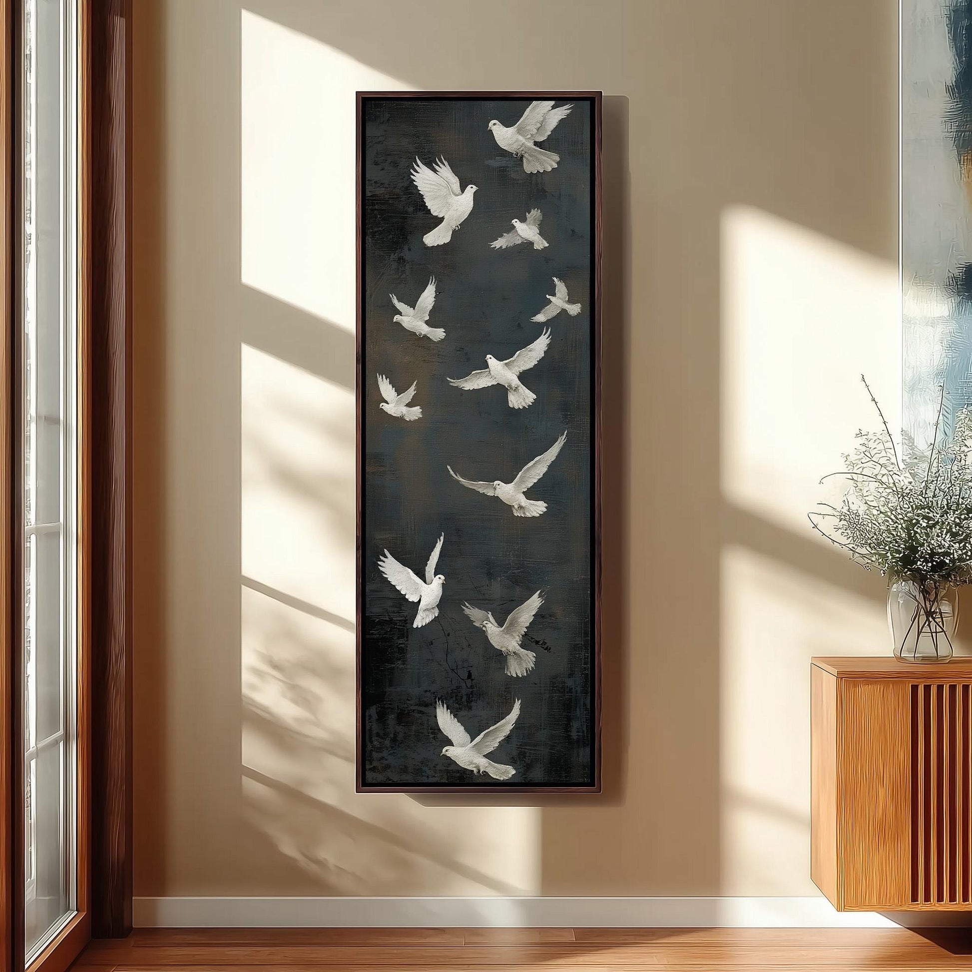 a large framed painting of white birds flying against a dark background, hanging on a wall in a room with a wooden cabinet and a vase of flowers.