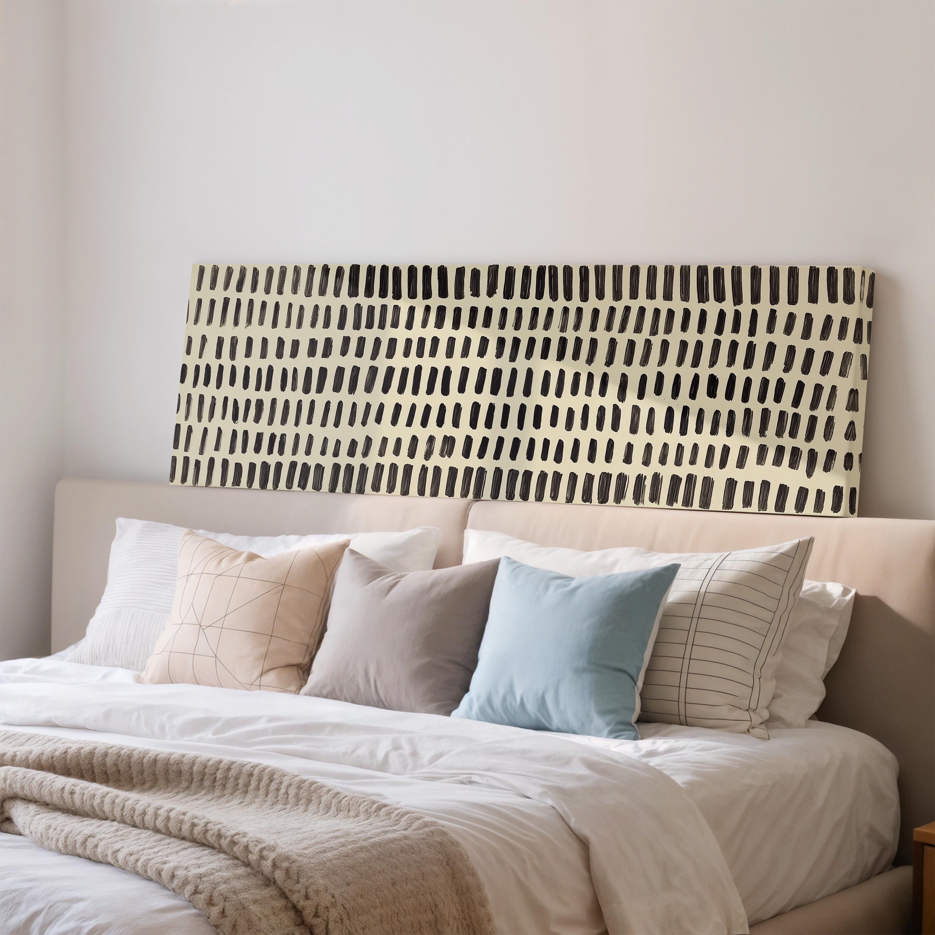 a neatly made bed with a white comforter and various pillows in different colors and patterns. The bed is positioned against a wall with a large abstract painting or artwork hanging above it.