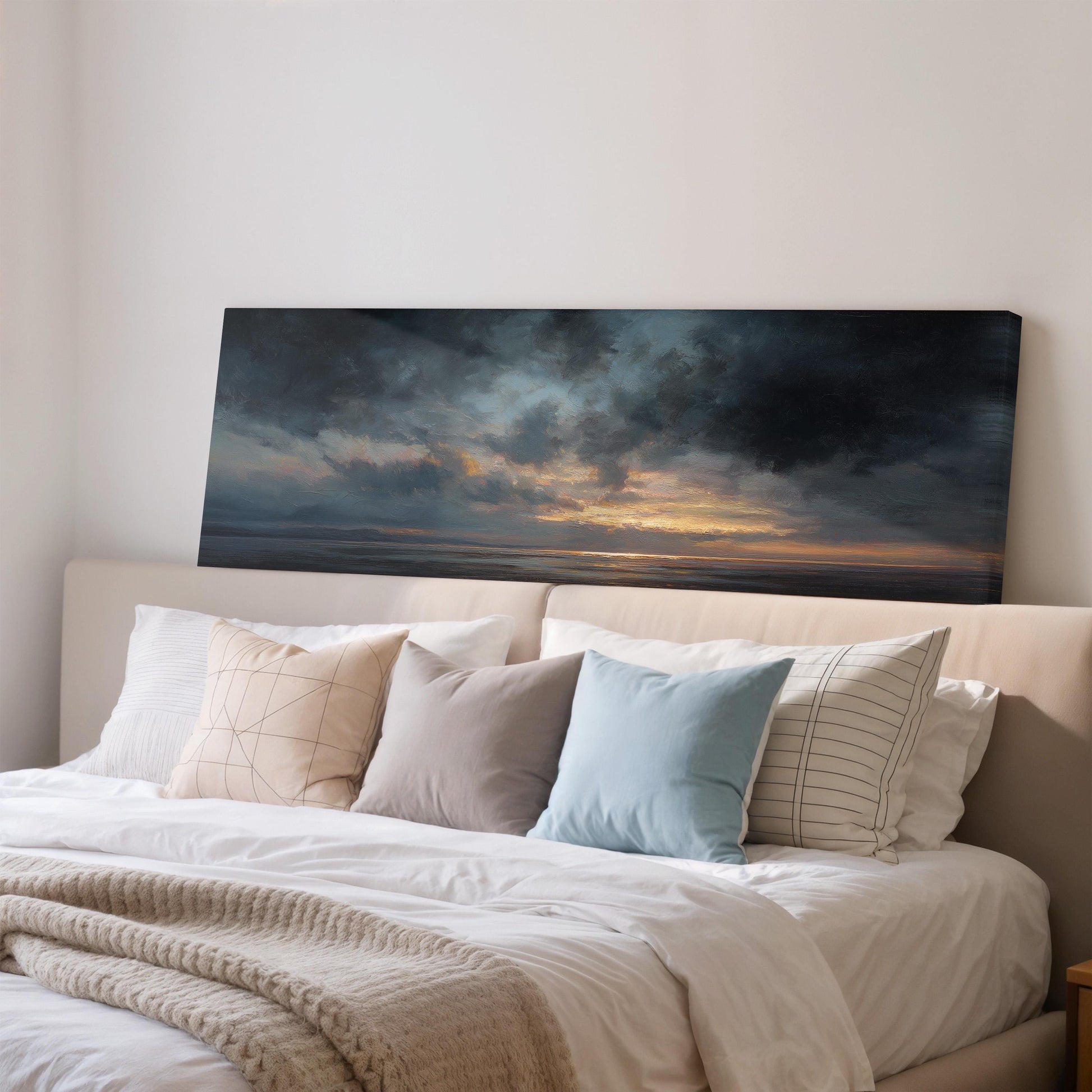 a large painting of a sunset over the ocean, hanging above a bed with white pillows and a gray blanket.