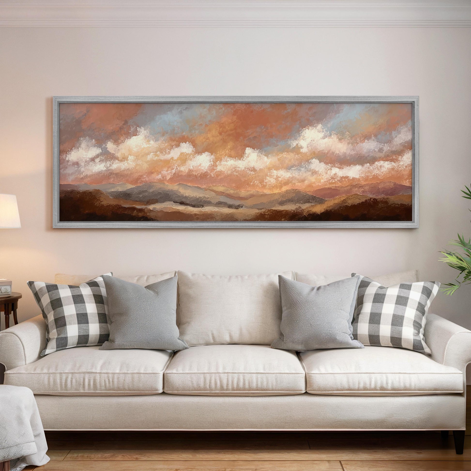 a large, framed painting of a landscape with mountains and clouds hanging on a wall above a white sofa with patterned throw pillows.