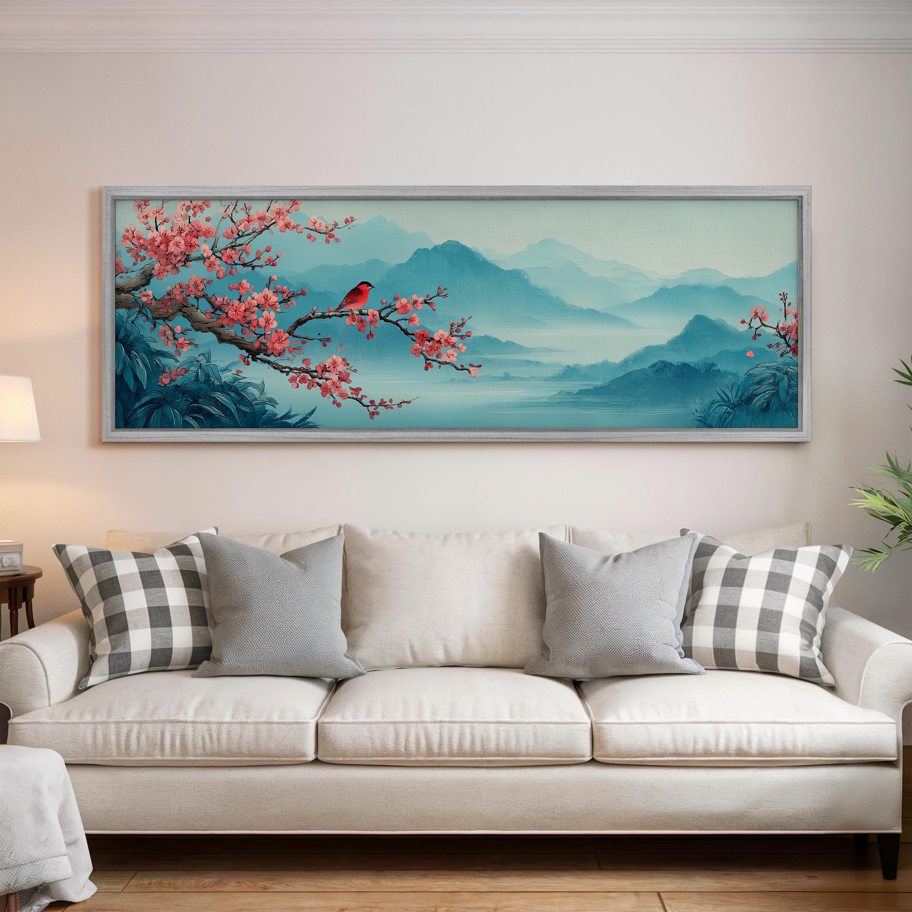 a large framed painting of a serene landscape with a bird perched on a branch, surrounded by mountains and trees. The painting is hung on a wall above a white couch with gray and white patterned pillows.