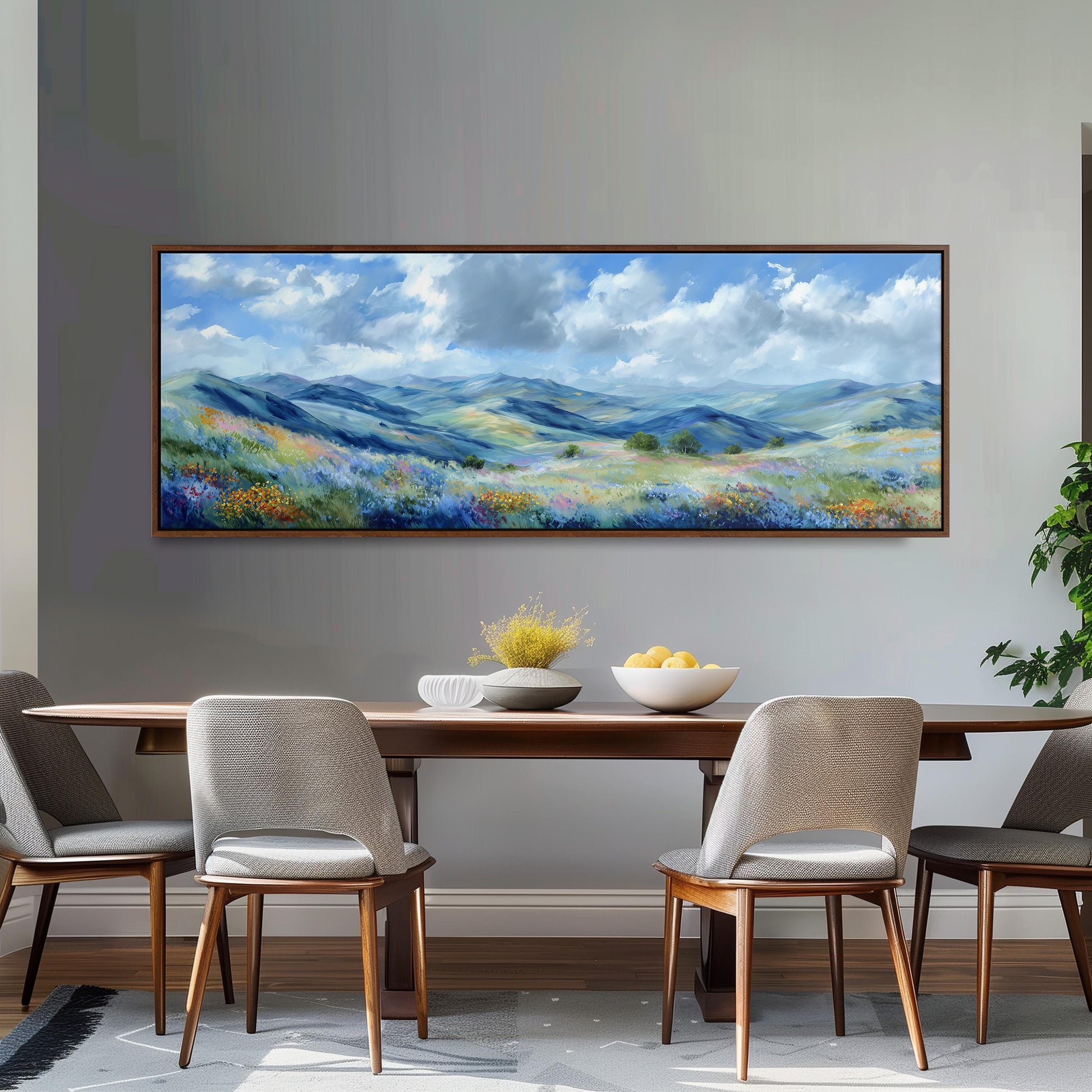 a large painting of a scenic landscape hanging on a wall above a wooden dining table with four chairs. The painting depicts a picturesque countryside scene with rolling hills, a blue sky with clouds, and a field of flowers. The dining table is set with a bowl of fruit and a vase of flowers, creating a cozy and inviting atmosphere.
