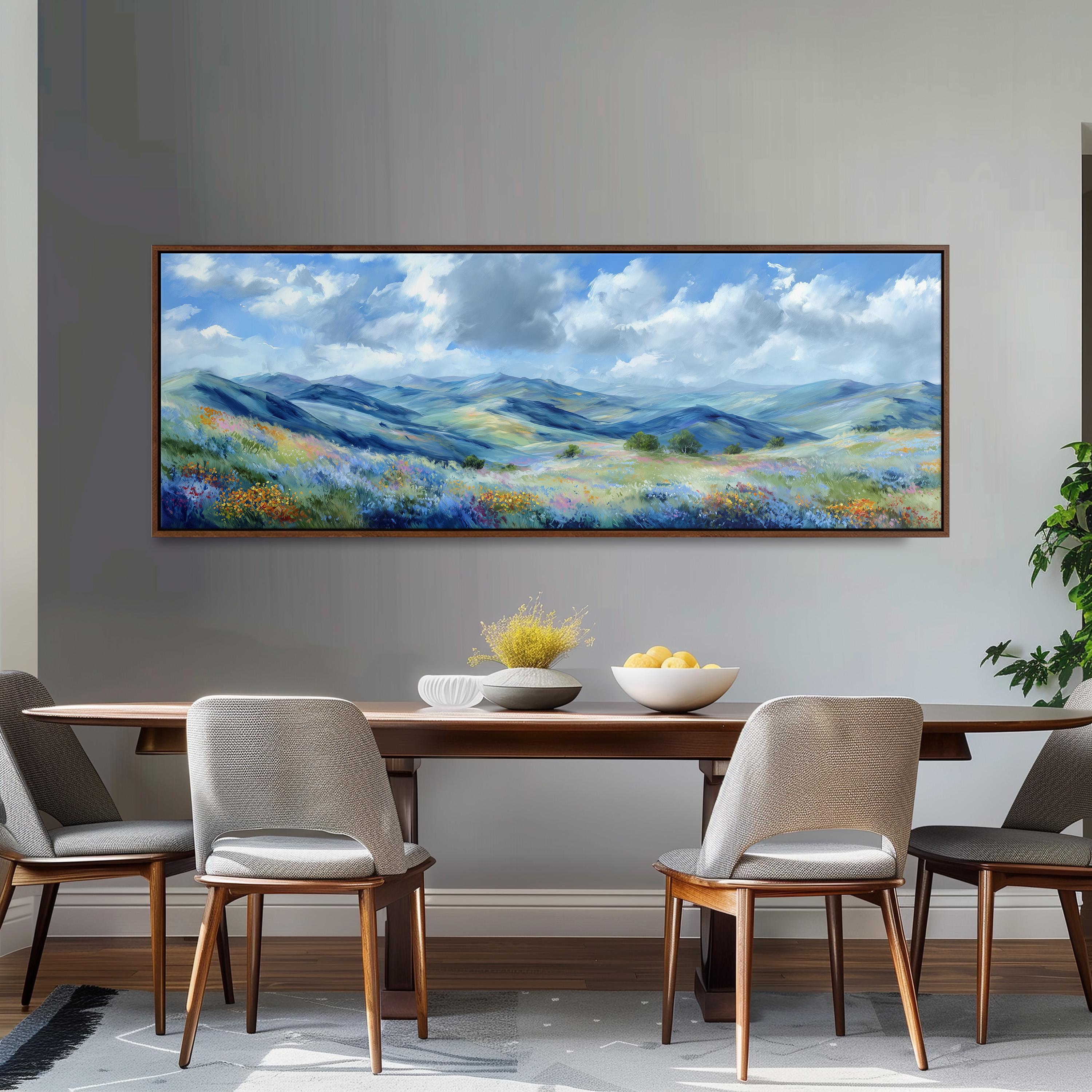 a large painting of a scenic landscape hanging on a wall above a wooden dining table with four chairs. The painting depicts a picturesque countryside scene with rolling hills, a blue sky with clouds, and a field of flowers. The dining table is set with a bowl of fruit and a vase of flowers, creating a cozy and inviting atmosphere.
