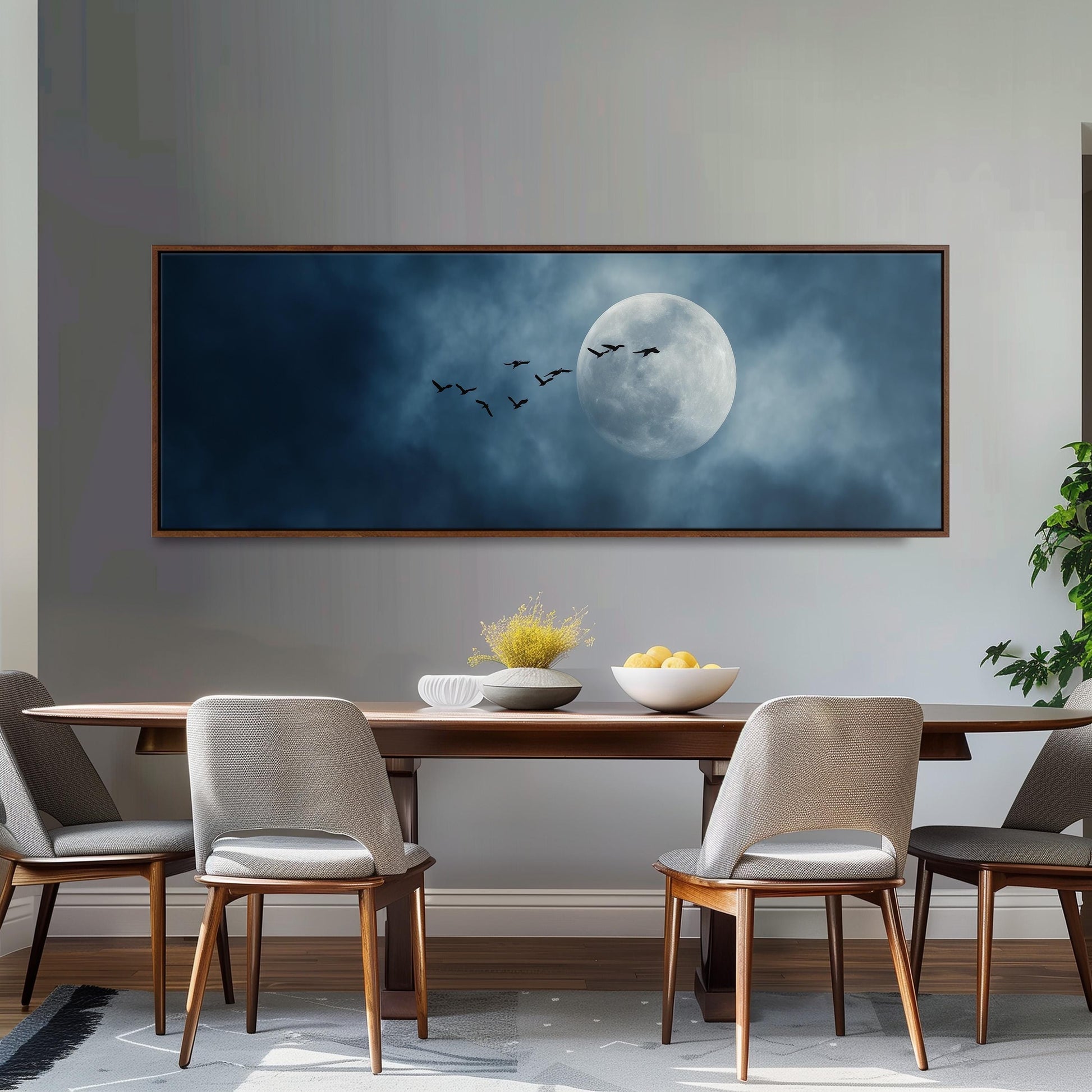 a large framed picture of a moonlit sky with birds flying across it, hanging on a wall above a wooden dining table with four chairs.