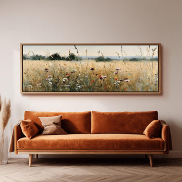 a large, framed painting of a field with tall grass and wildflowers hanging on a wall above a brown sofa.