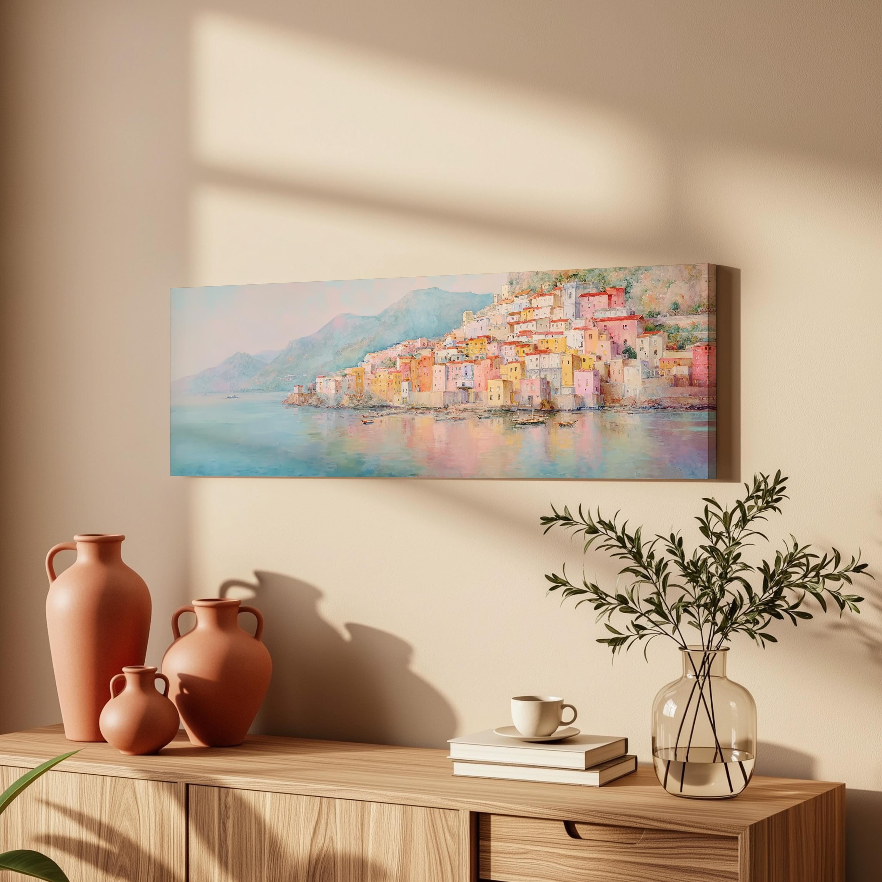 a wooden cabinet with a painting of a colorful coastal town hanging on the wall above it. The cabinet also has three orange vases and a glass vase with greenery, as well as a stack of books and a cup on the surface.
