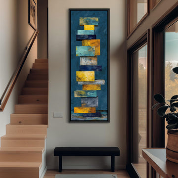 a modern art piece hanging on the wall, depicting a stack of colorful geometric shapes in shades of blue, yellow, and orange. The artwork is framed and positioned above a black bench, which is located in the corner of the room. The room has a wooden staircase leading up to the second floor, and a window can be seen on the right side of the image.