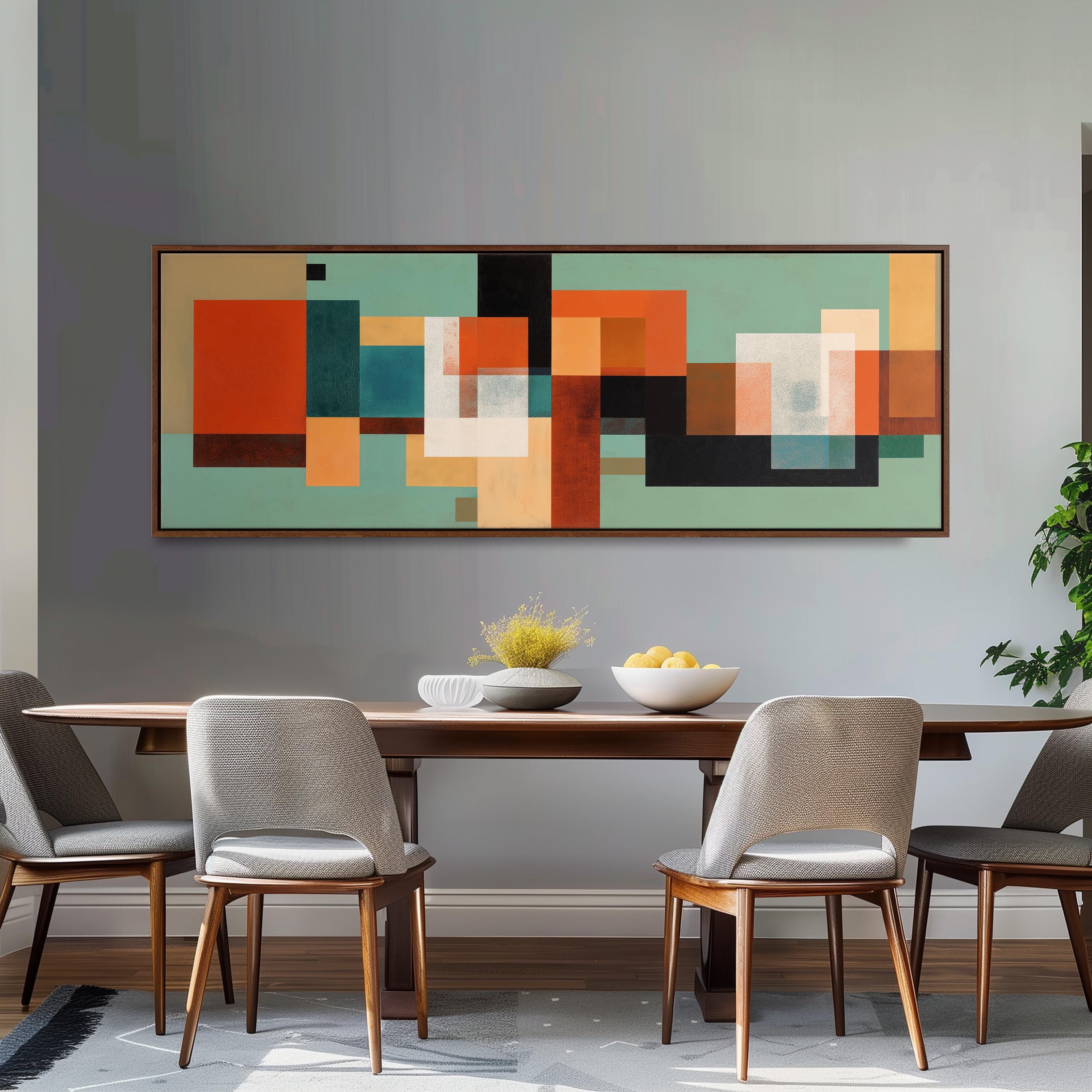 a modern dining room with a wooden table and four gray chairs. Above the table, there is a large abstract painting with a geometric design in shades of green, orange, and black. The table is set with a bowl of fruit and a vase of flowers, creating a welcoming and stylish atmosphere.