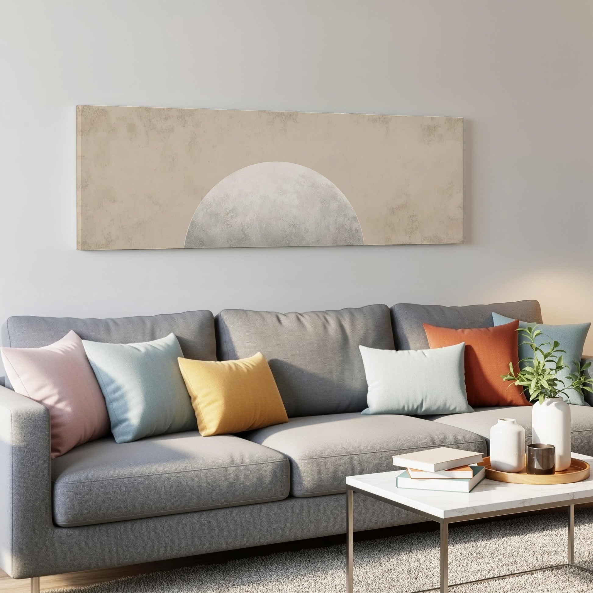 a modern living room with a gray sofa adorned with colorful throw pillows. Above the sofa, there is a large rectangular artwork depicting a crescent moon. In front of the sofa, there is a white coffee table with a stack of books and a small plant.