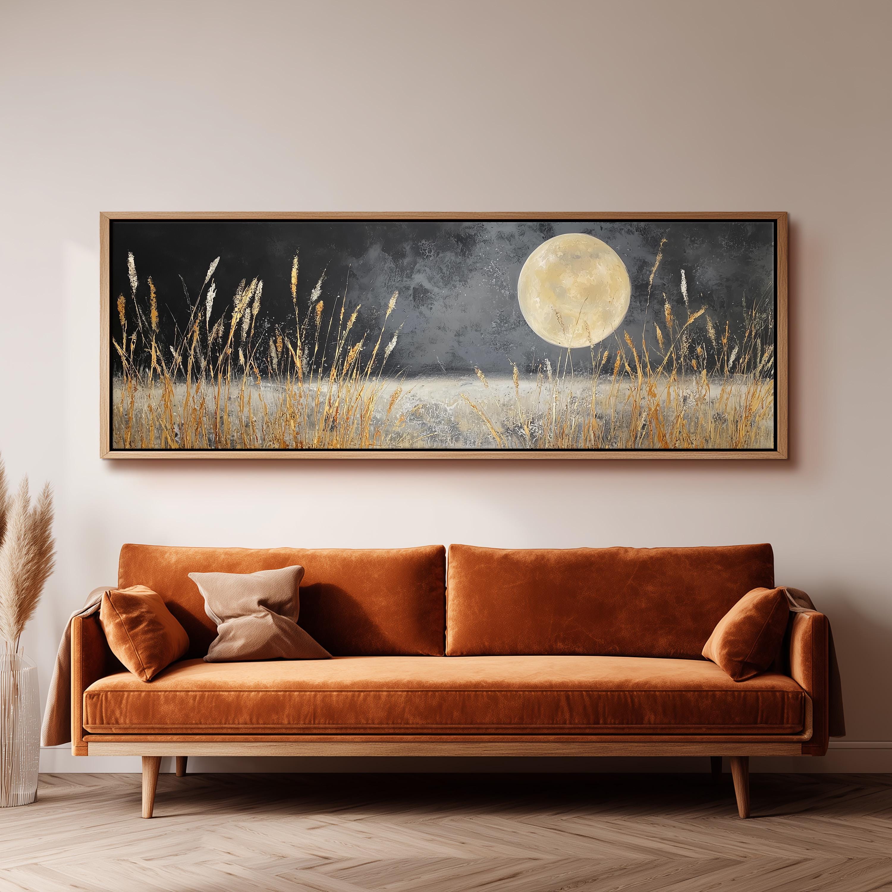 a large, framed painting of a field with tall grass and a full moon in the night sky, hanging on a wall above a brown sofa.