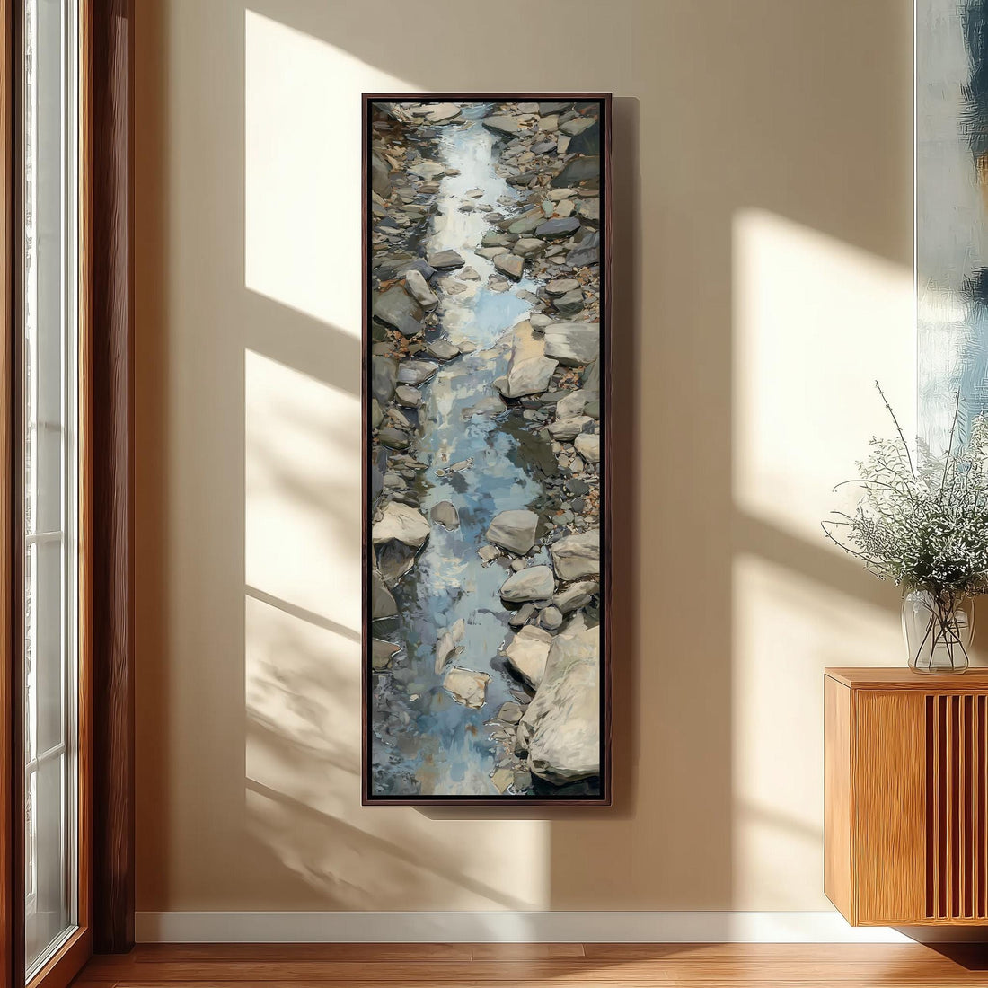 a large framed painting of a rocky stream or river, hanging on a wall in a room with a wooden cabinet and a vase of flowers.
