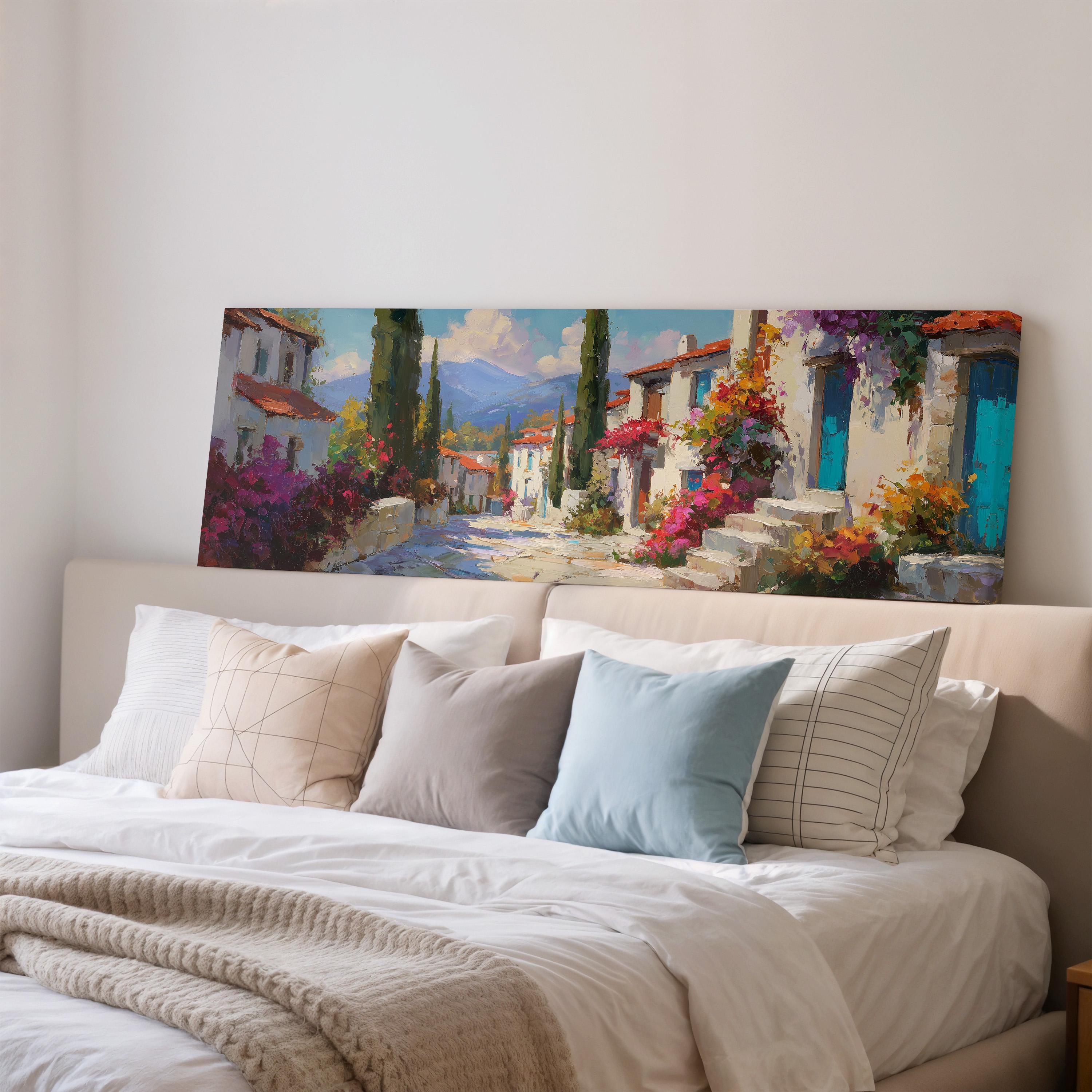 a large painting of a picturesque village scene hanging on a wall above a bed with white pillows and a gray blanket.