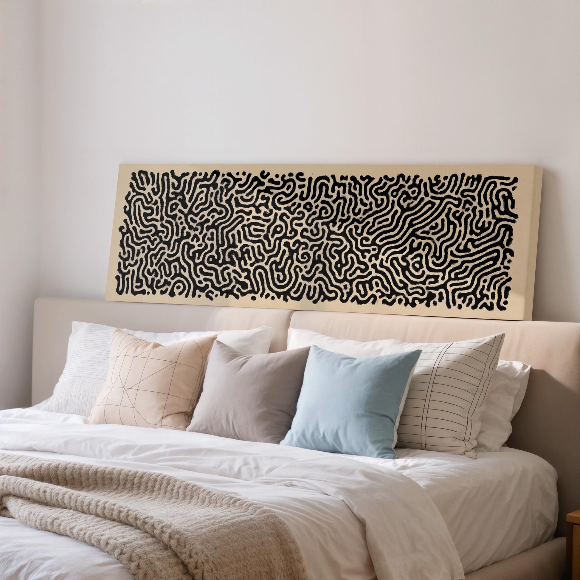 a large bed with a white comforter and a variety of pillows, including a blue pillow and a striped pillow. Above the bed, there is a large abstract painting or artwork with a black and white color scheme.