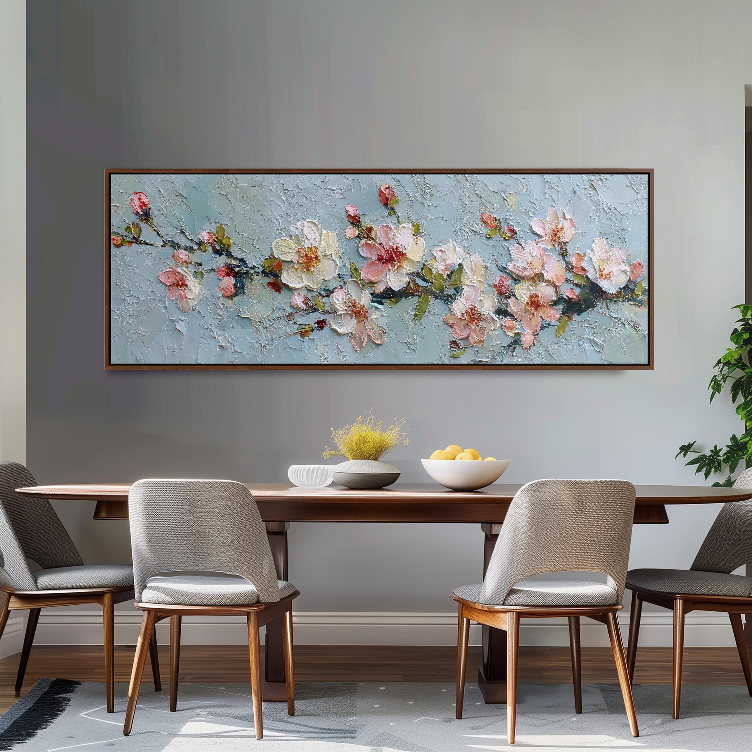 a dining room with a large painting of pink flowers hanging on the wall above a wooden table with four chairs. The table is set with a bowl of fruit and a vase of flowers, creating a cozy and inviting atmosphere.