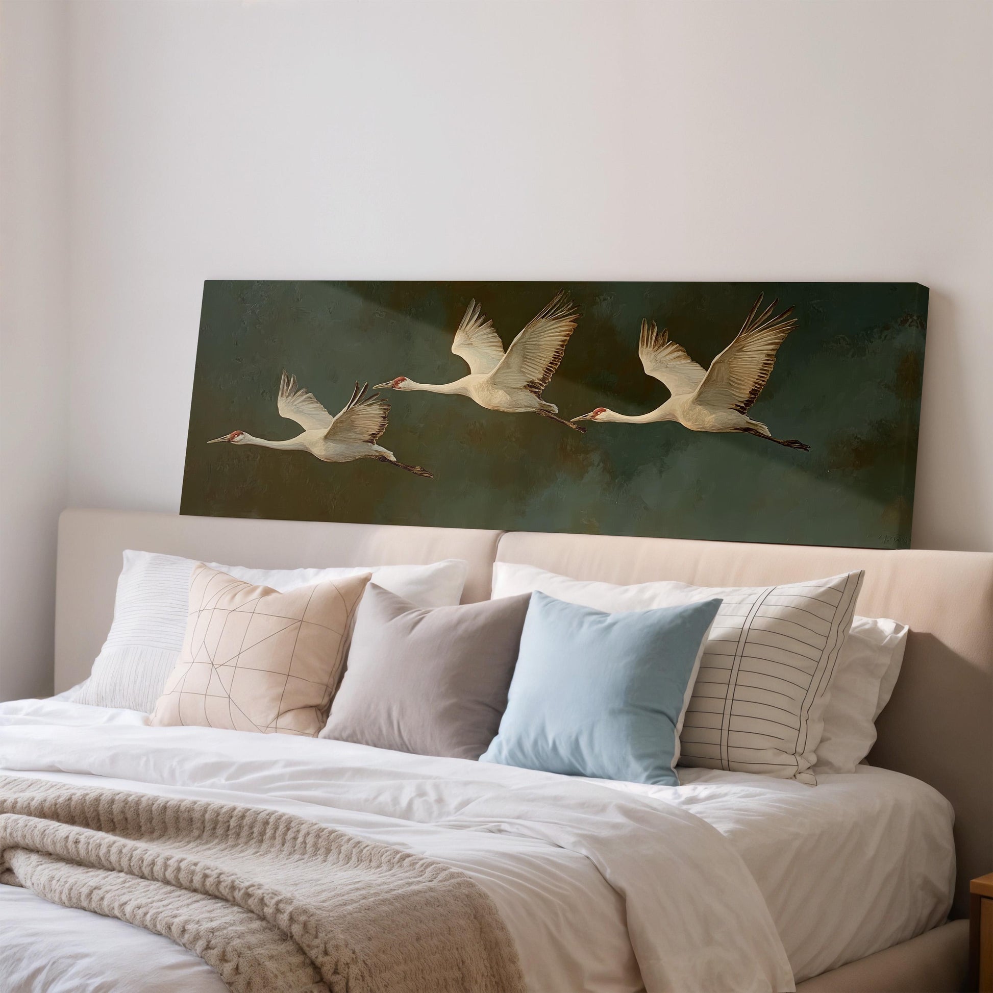 a large painting of three white birds in flight on a dark green background, displayed on a wall above a bed with white pillows and a gray blanket.