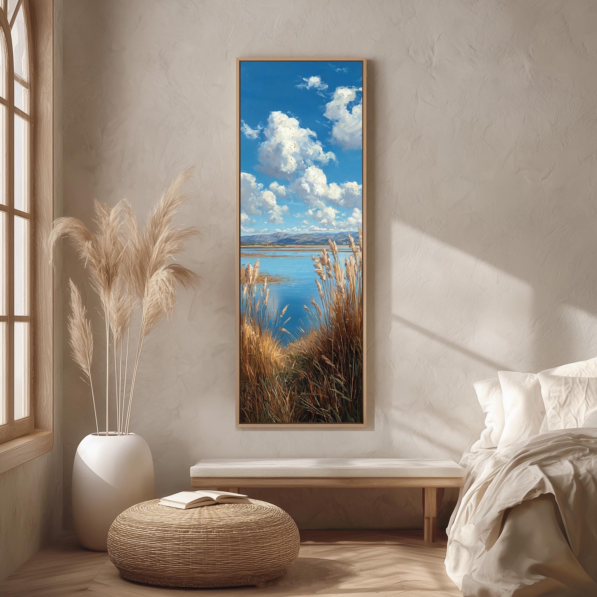 a serene bedroom scene with a large framed painting of a tranquil landscape featuring a blue sky, fluffy white clouds, and a body of water. The painting is hung on a wall above a wooden bench, and the room has a minimalist and modern design with a white color scheme and natural elements like a potted plant and a woven ottoman.