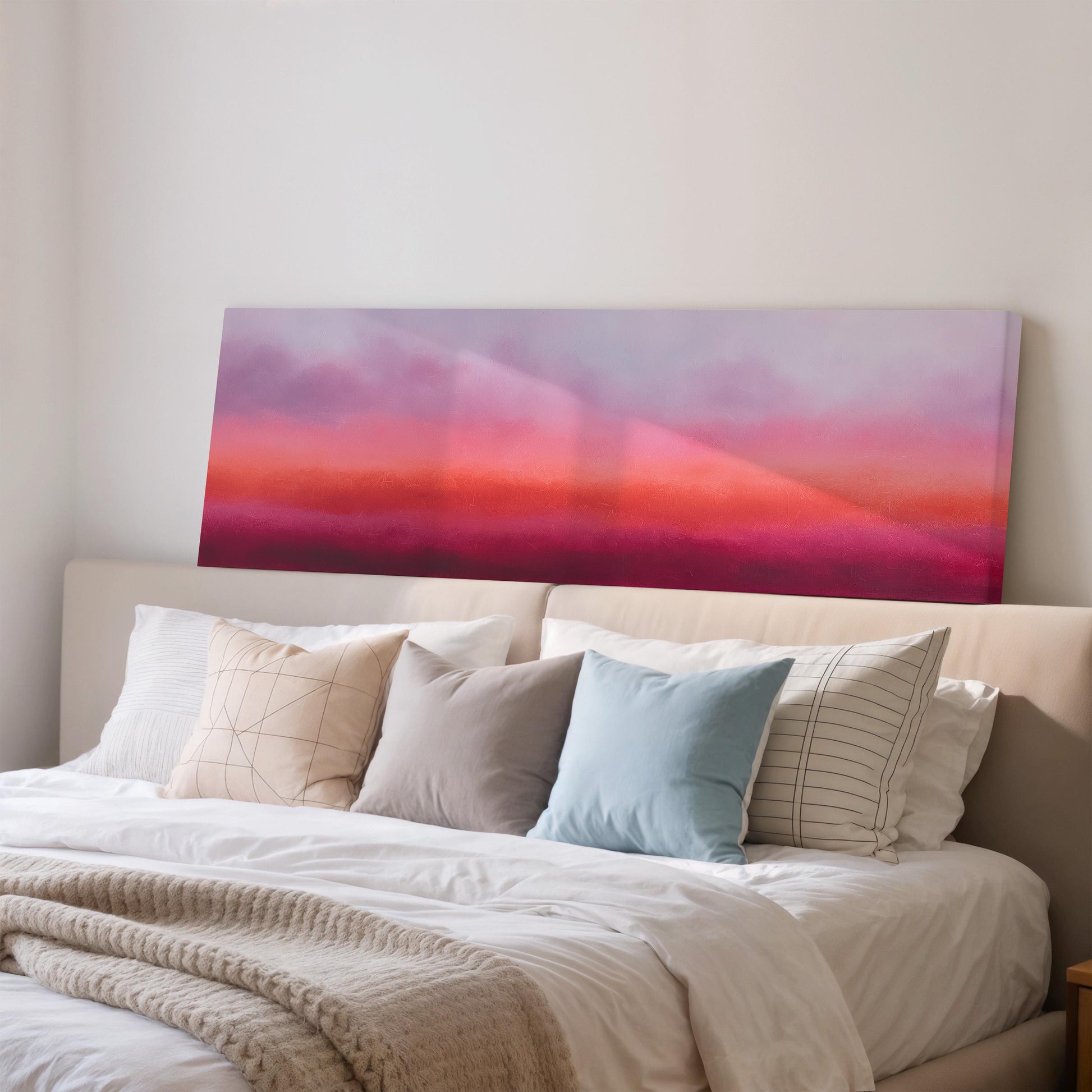 a cozy bedroom with a large bed adorned with white linens and a variety of colorful pillows. Above the bed, there is a large canvas print depicting a vibrant sunset scene in shades of pink and purple.